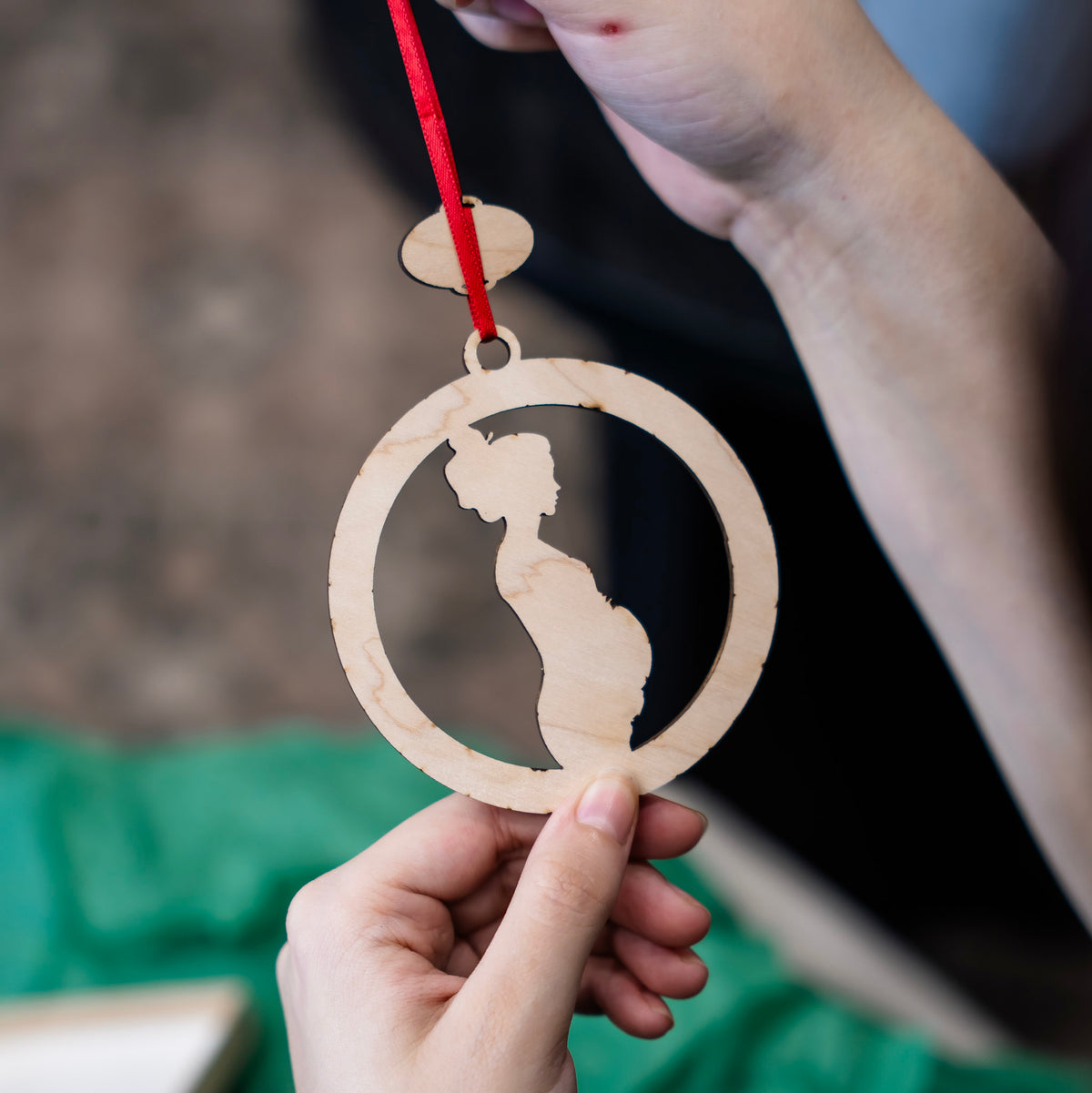 A close-up of a hand holding the backside of a personalized wooden baby bump ornament with a red ribbon. The ornament features a laser-cut silhouette of a pregnant belly