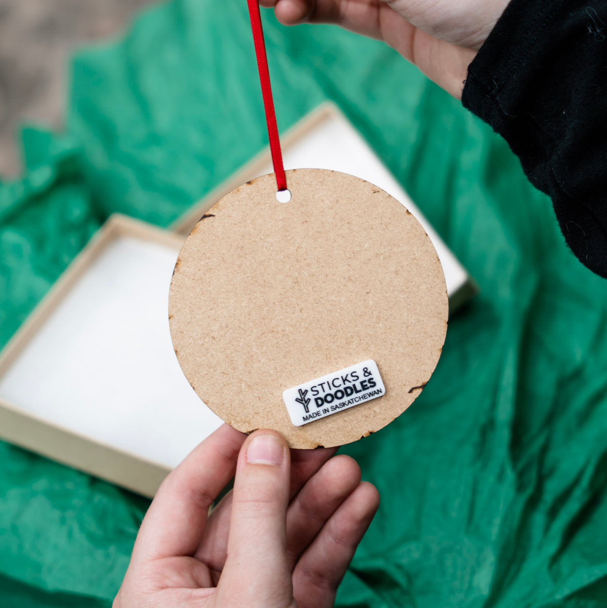 A person holds the back of a round wood ornament with a tag that reads STICKS AND DOODLES MADE IN SASKATCHEWAN. There is a light wood box and crinkled green tissue paper are visible in the background.