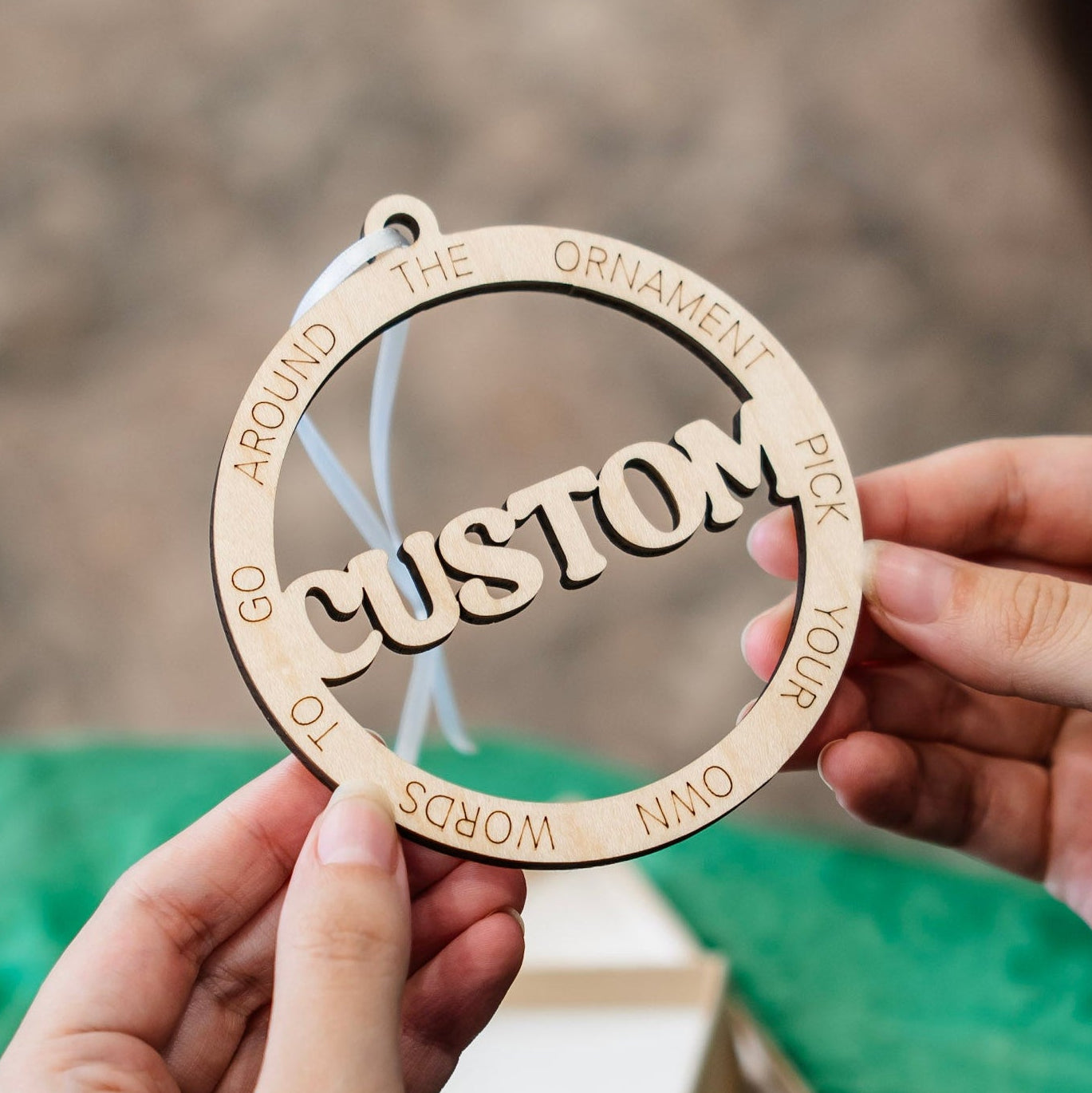 A person holds a round wooden ornament with the word 'CUSTOM' in the center and engraved words around the edge, against a softly blurred outdoor background with green wrapping paper visible.
