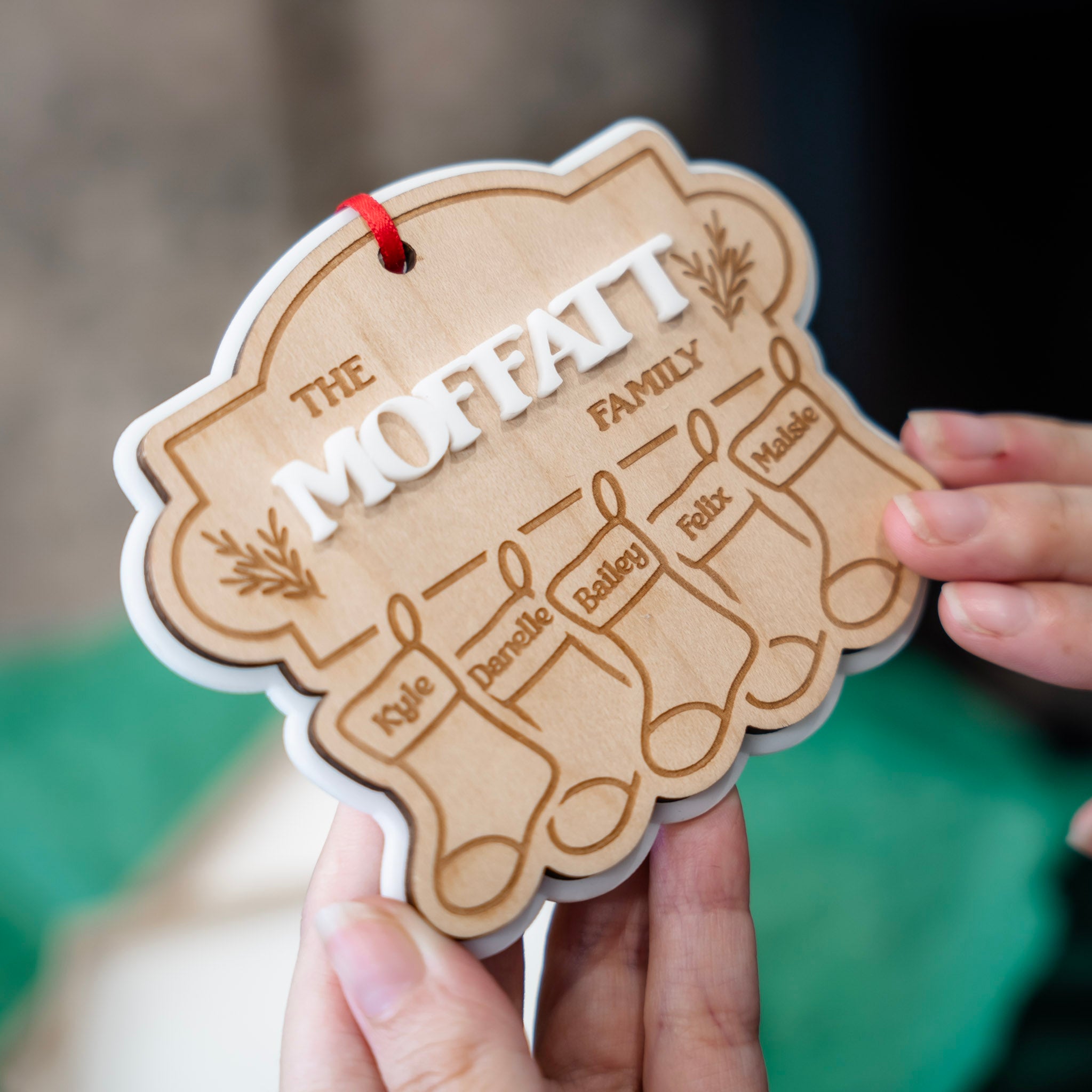 A close-up of a hand holding a personalized wooden Christmas ornament shaped like stockings, engraved with the name 'The Moffatt Family' and individual names for each stocking, with a red hanging ribbon.