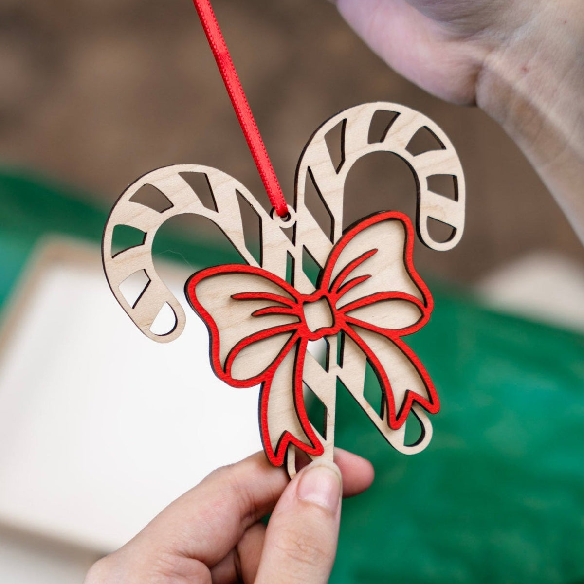 A close-up of hands holding a laser-cut wooden Christmas ornament shaped like two candy canes with a red bow, suspended by a red ribbon against a soft-focus background of green tissue paper and a gift box.