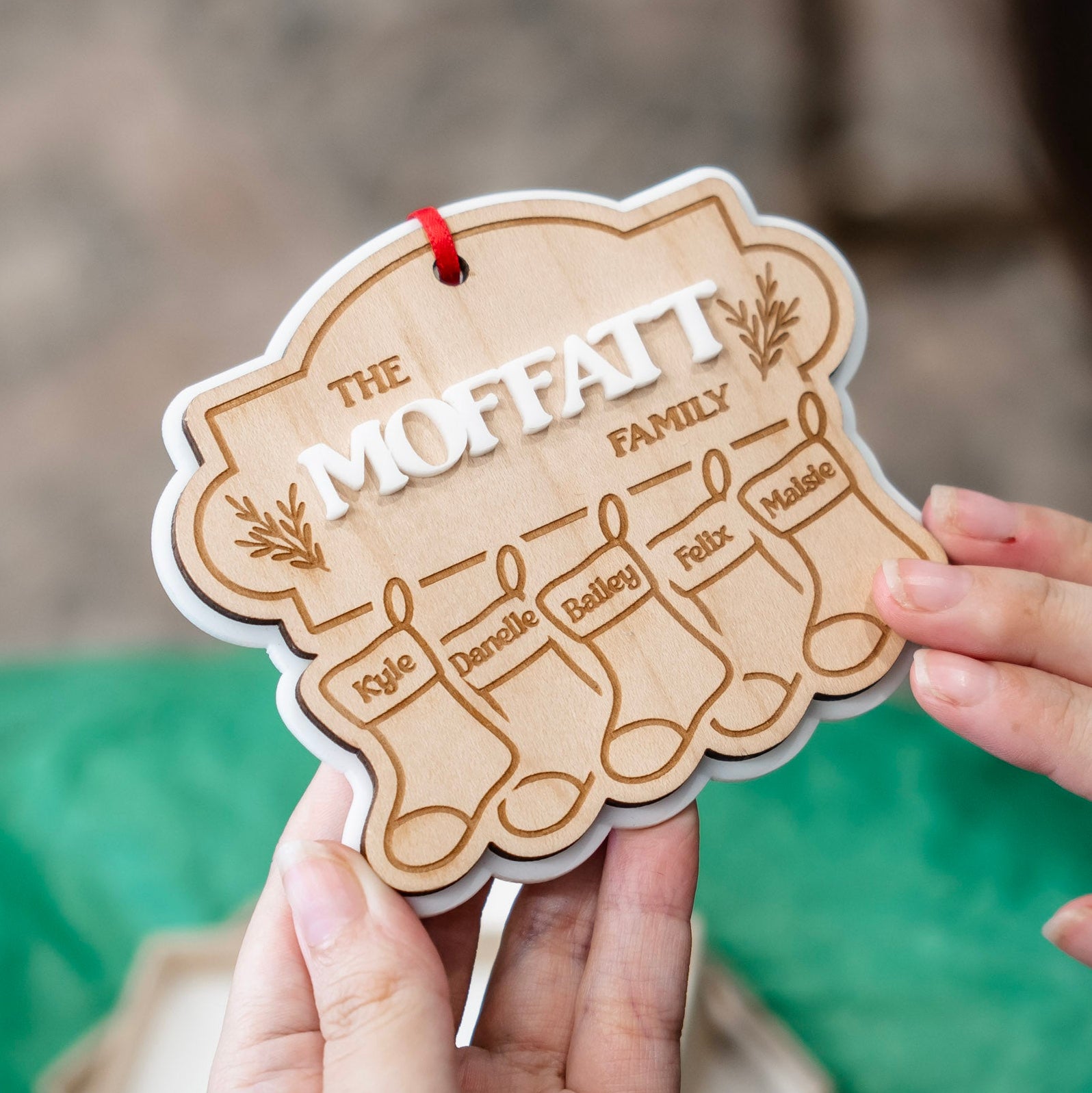 A personalized wooden Christmas ornament shaped like four stockings, held by hands against a blurred background with a green fabric surface.
