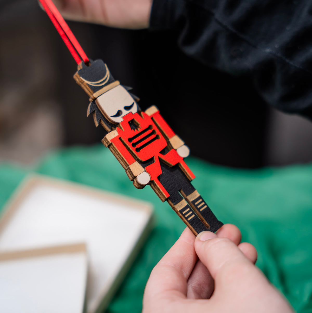 A close-up of a hand holding a wooden nutcracker ornament with a red ribbon, against a soft-focus background with green tissue paper and a gift box.
