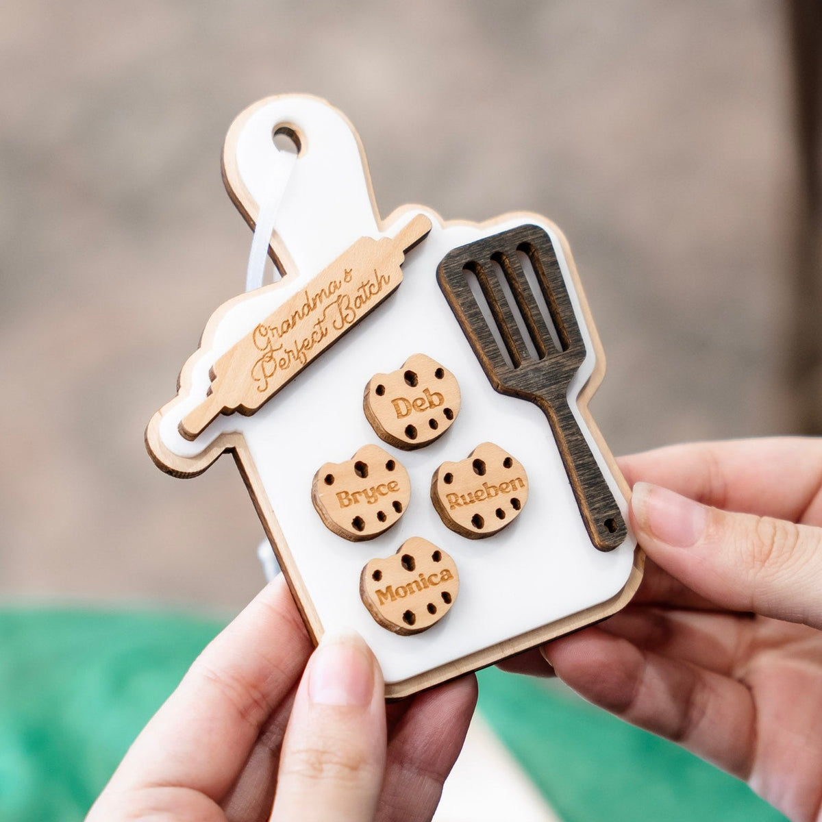 A personalized wooden baking-themed keepsake ornament held in two hands. The ornament is shaped like a white baking tray with a wooden rim, featuring a wooden rolling pin, a dark wooden spatula, and four round wooden cookie cutouts engraved with names: Deb, Bryce, Rueben, and Monica.
