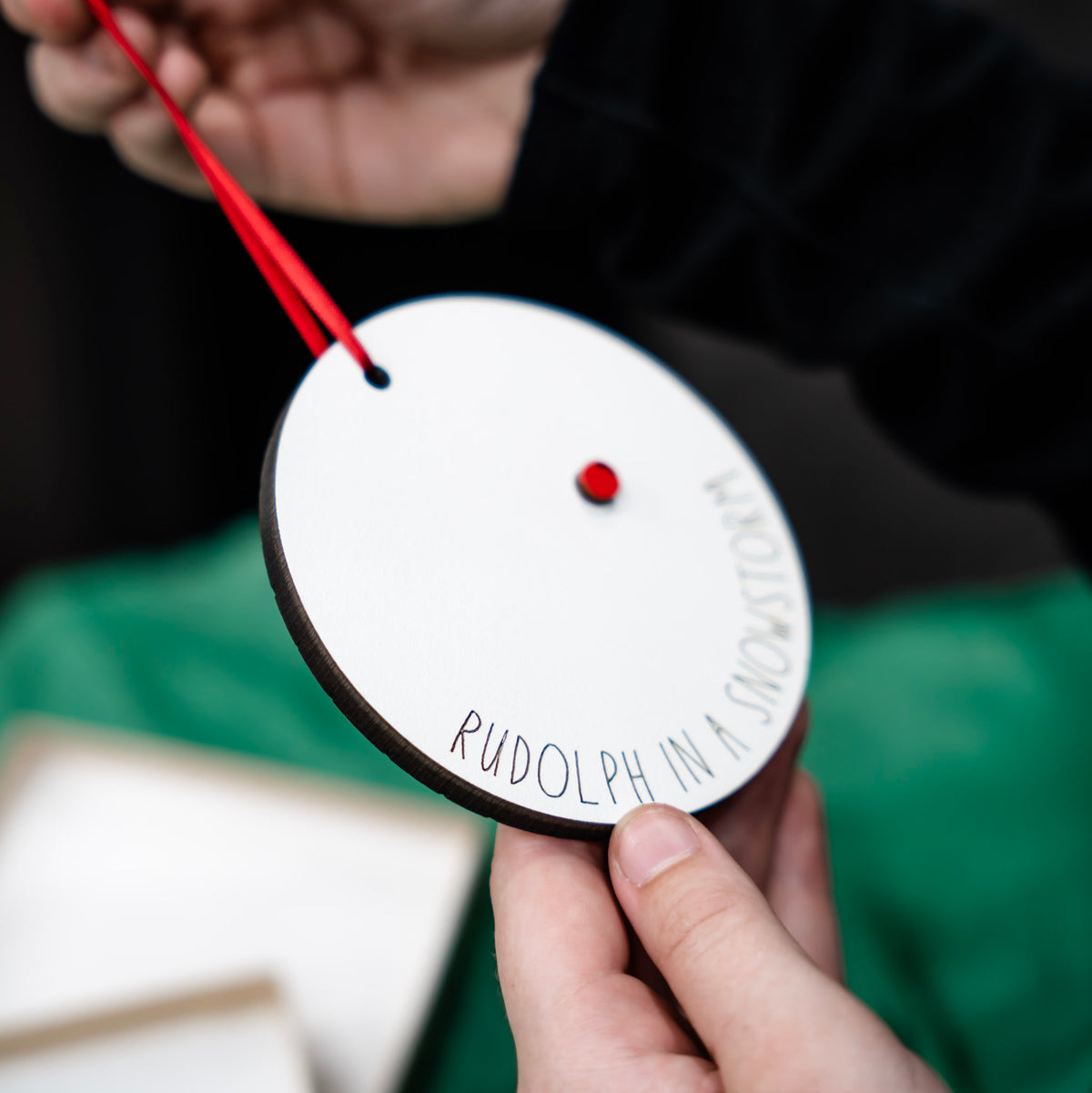 A side close up view of a round white ornament with a red hanging ribbon and a small red dot, engraved with the text “RUDOLPH IN A SNOWSTORM”. A light wood box and crinkled green tissue paper are visible in the background.