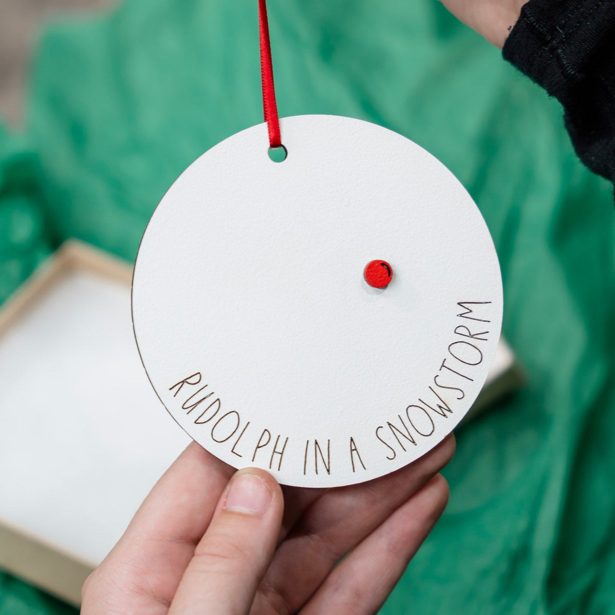 A person holds a round white ornament with a red hanging ribbon and a small red dot, engraved with the text “RUDOLPH IN A SNOWSTORM”. A light wood box and crinkled green tissue paper are visible in the background.