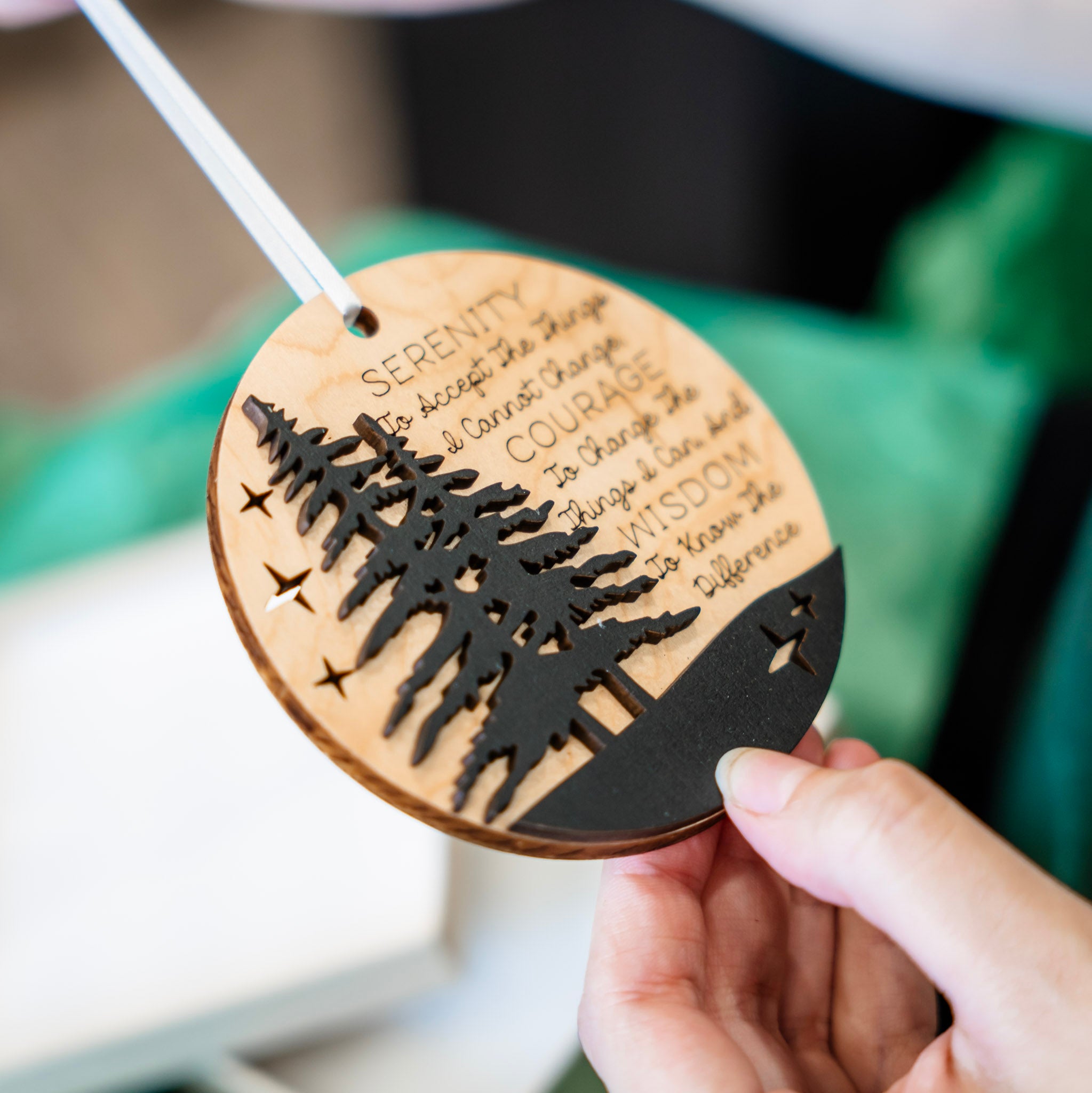 A person holds a round wooden ornament with a laser-cut tree silhouette and engraved inspirational quote. The ornament has a natural light wood finish with a black lower band and black cutout details, suspended by a white ribbon.