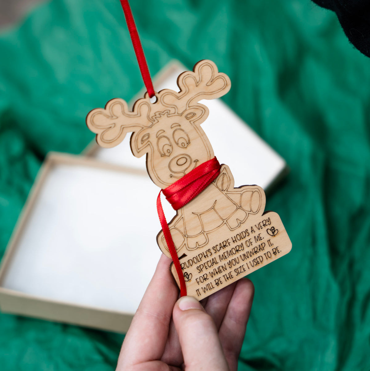 A hand holds a laser-engraved wooden Christmas ornament shaped like a reindeer, wrapped with a red ribbon that can be cut to the right length, with a red hanging red ribbon. The ornament features engraved text.
