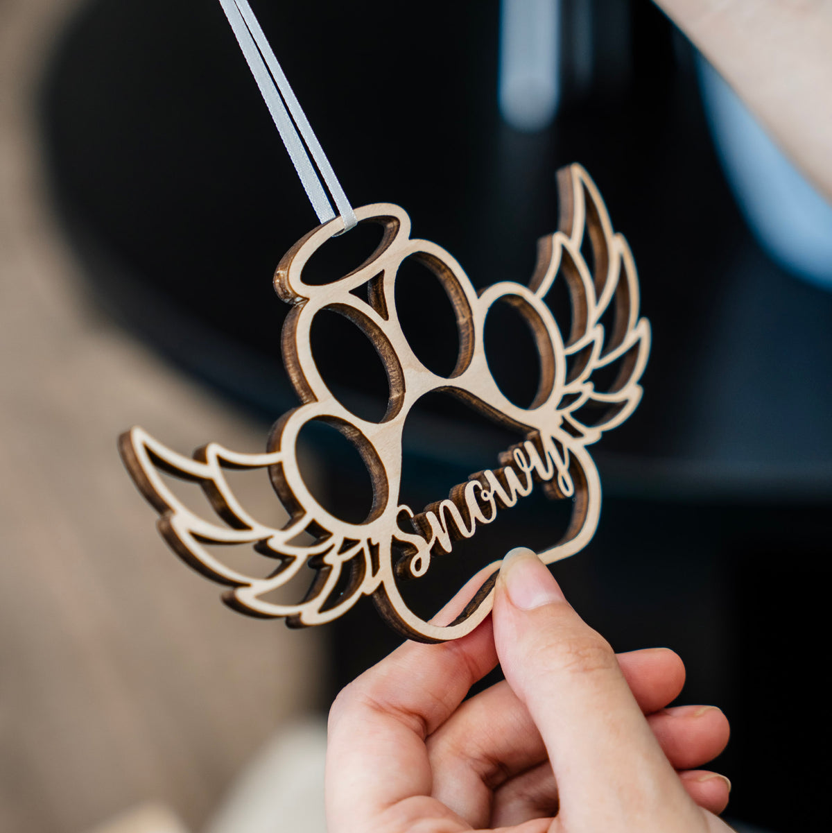 A hand holding a personalized wooden paw print ornament with angel wings and the name 'Snowy' cut into it, shown against a dark, blurred background.