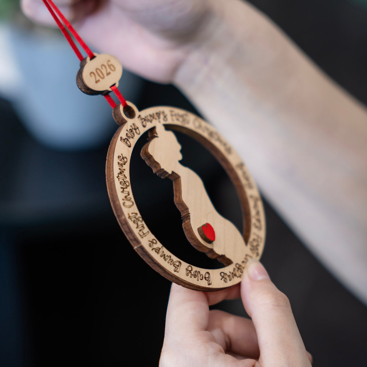 A close-up of a hand holding a personalized wooden baby bump ornament with a red ribbon. The ornament features a laser-cut silhouette of a pregnant belly with a small red heart, surrounded by engraved phrases, with a small wooden tag at the top engraved with '2026'.