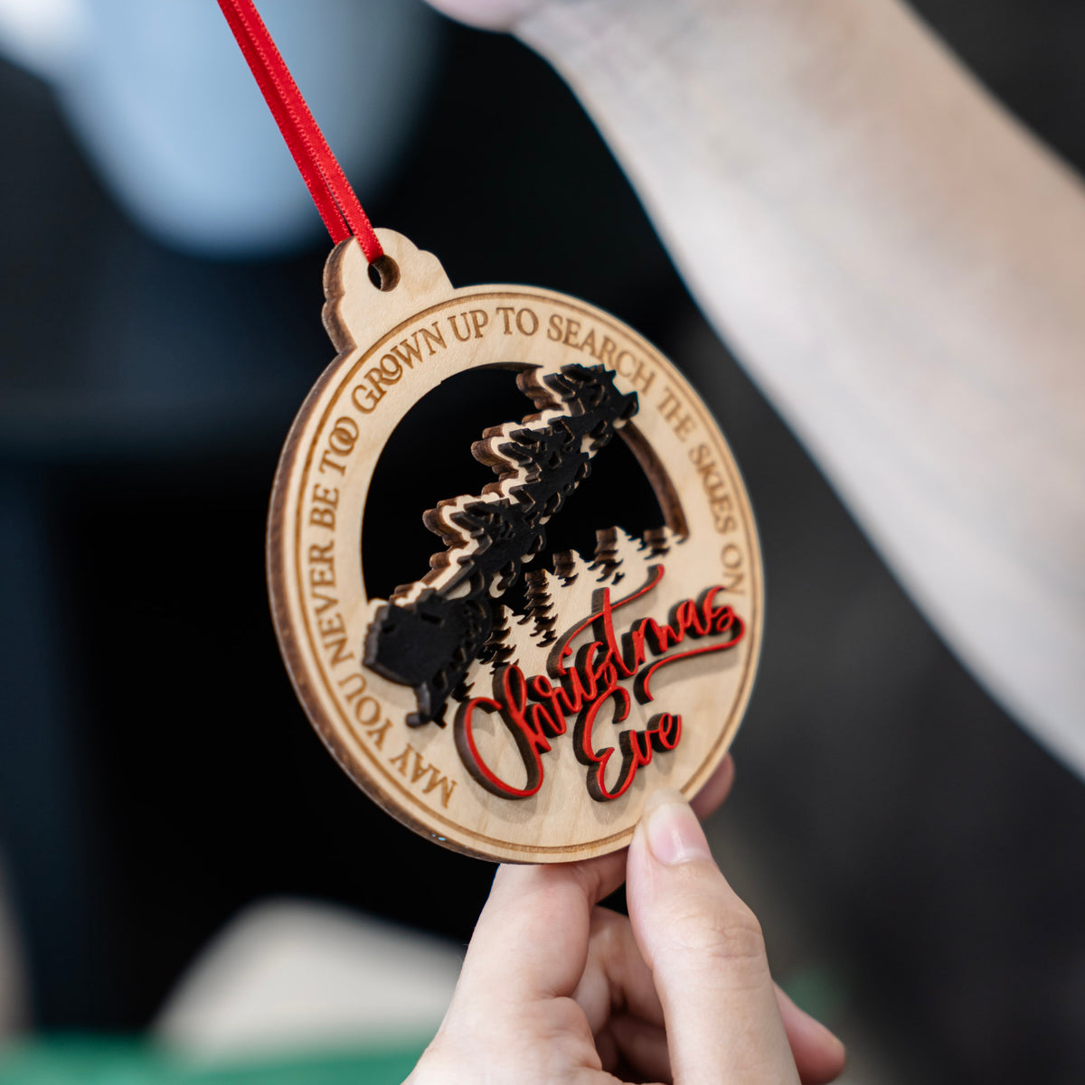 A close up of a laser-engraved wooden Christmas Eve ornament held by hands, with a red ribbon, Santa's sleigh silhouette, pine trees, and the text 'May you never be too grown up to search the seas on Christmas Eve'.
