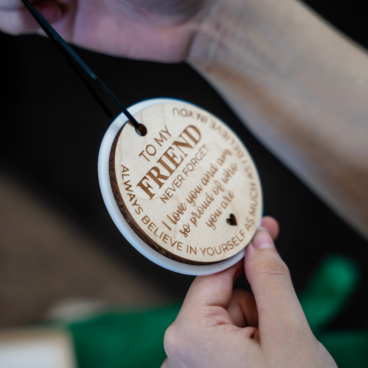 A close-up of a person holding a round wooden engraved ornament. The ornament features engraved text reading 'TO MY FRIEND NEVER FORGET I love you and am so proud of who you are. ALWAYS BELIEVE IN YOURSELF AS MUCH AS I BELIEVE IN YOU'.