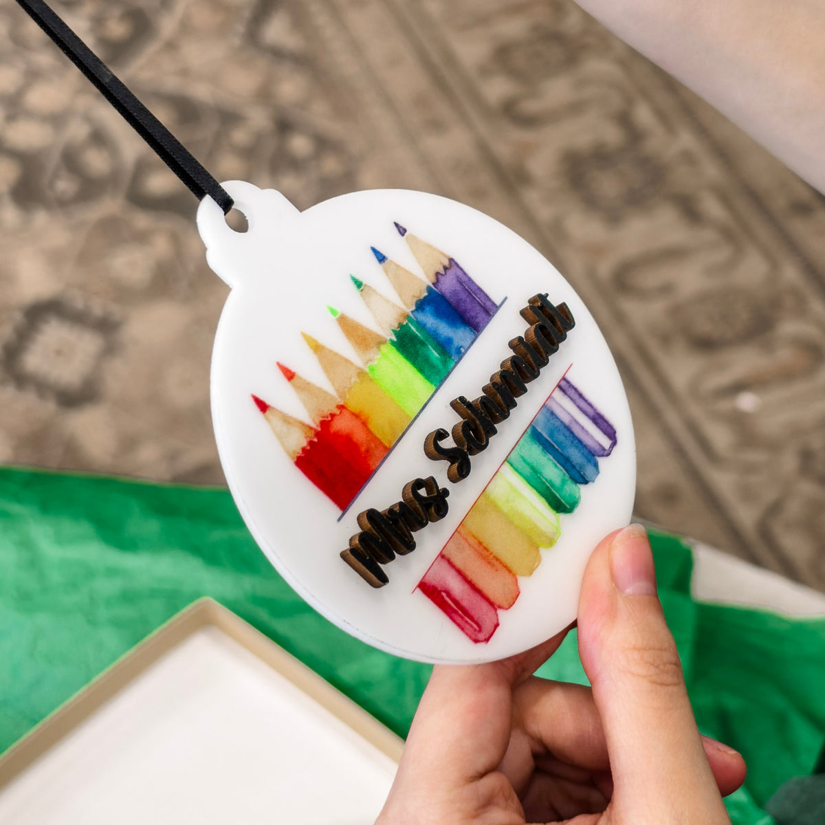 A hand holds a round white ornament featuring colorful pencil illustrations and the name 'Mrs. Schmidt' in black script, with a black hanging ribbon above it.