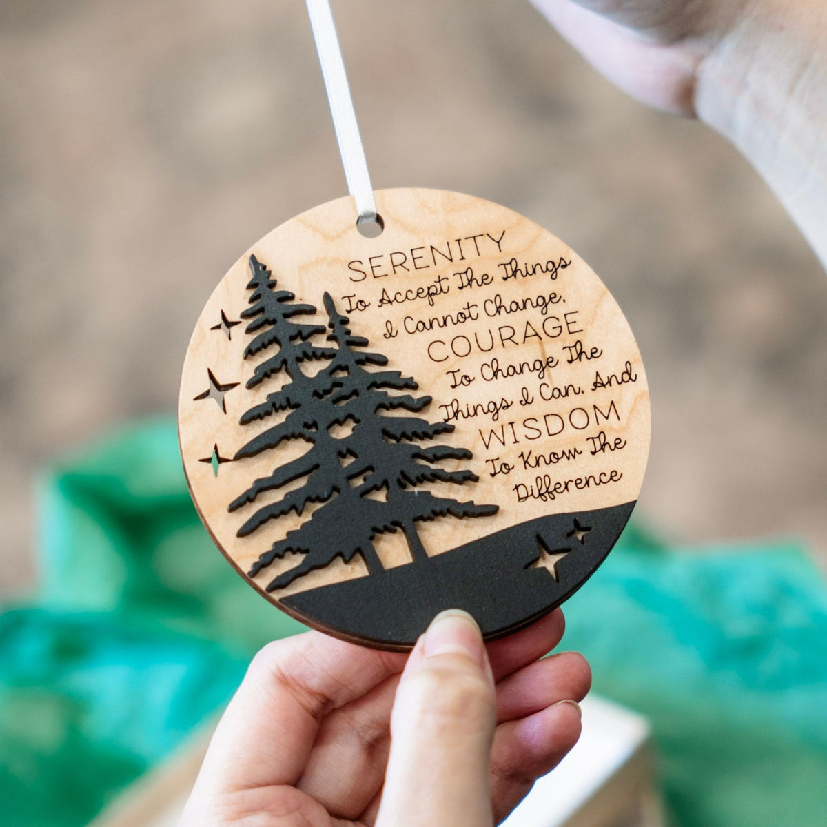 A person holds a round wooden ornament with a laser-cut tree silhouette and engraved inspirational quote. The ornament has a natural light wood finish with a black lower band and black cutout details, suspended by a white ribbon.