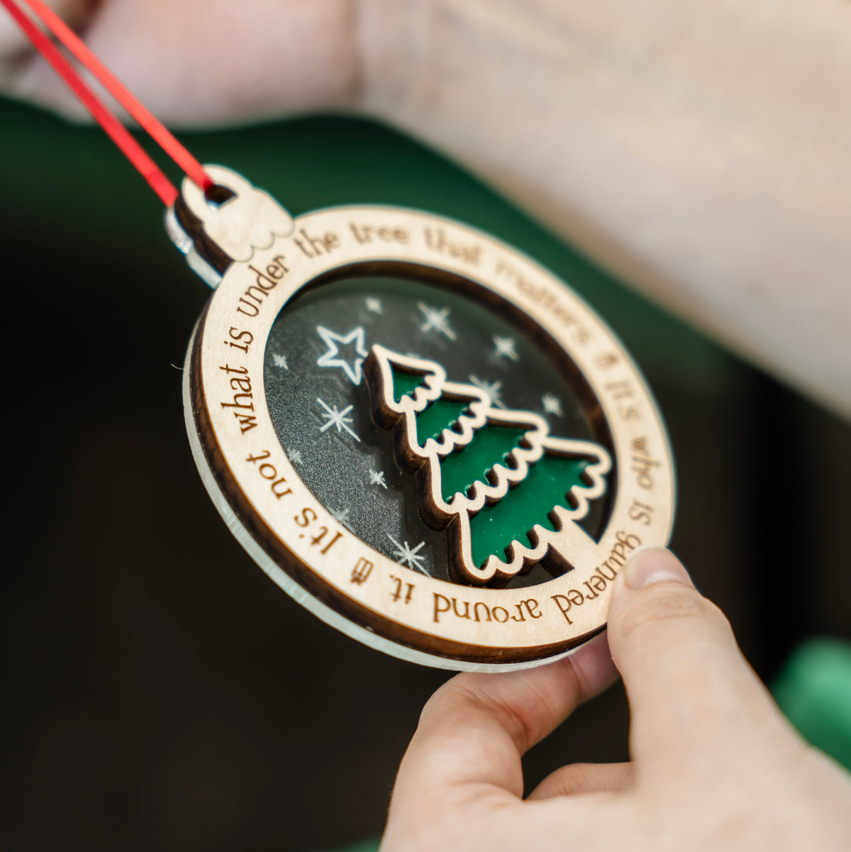 A person holds a round wooden Christmas ornament with a red hanging string. The ornament features a green Christmas tree ontop of a frosted acrylic backer that has stars scored onto it, and engraved text around the edge that says 'It's not what is under the tree that matters. It's who is gathered around it.', shown against a blurred background.