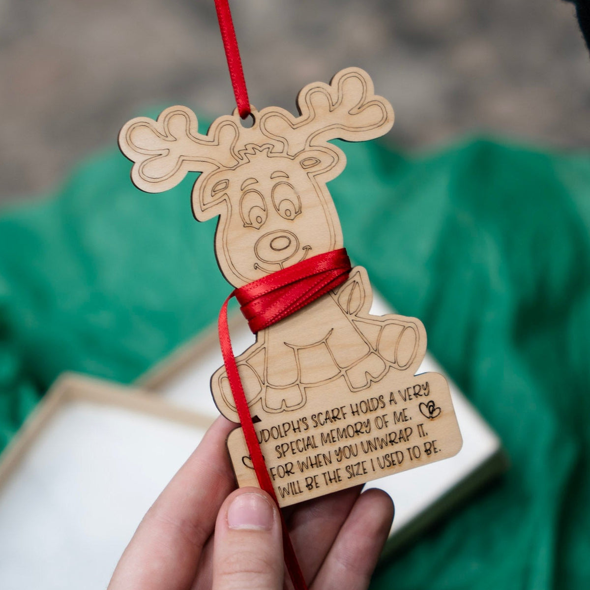 A hand holds a laser-engraved wooden Christmas ornament shaped like a reindeer, wrapped with a red ribbon that can be cut to the right length, with a red hanging red ribbon. The ornament features engraved text.