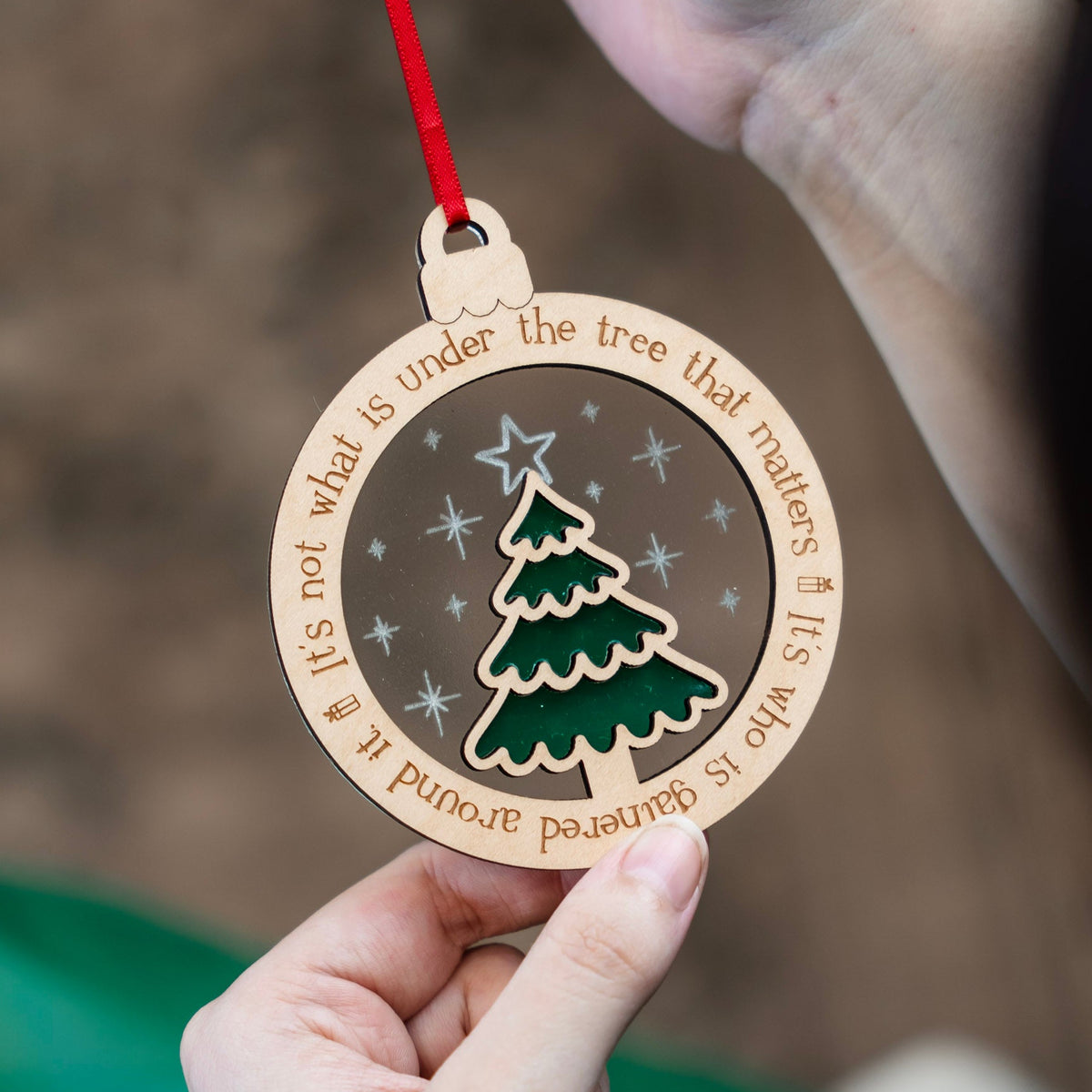 A person holds a round wooden Christmas ornament with a red hanging string. The ornament features a green Christmas tree ontop of a frosted acrylic backer that has stars scored onto it, and engraved text around the edge that says 'It's not what is under the tree that matters. It's who is gathered around it.', shown against a blurred background.