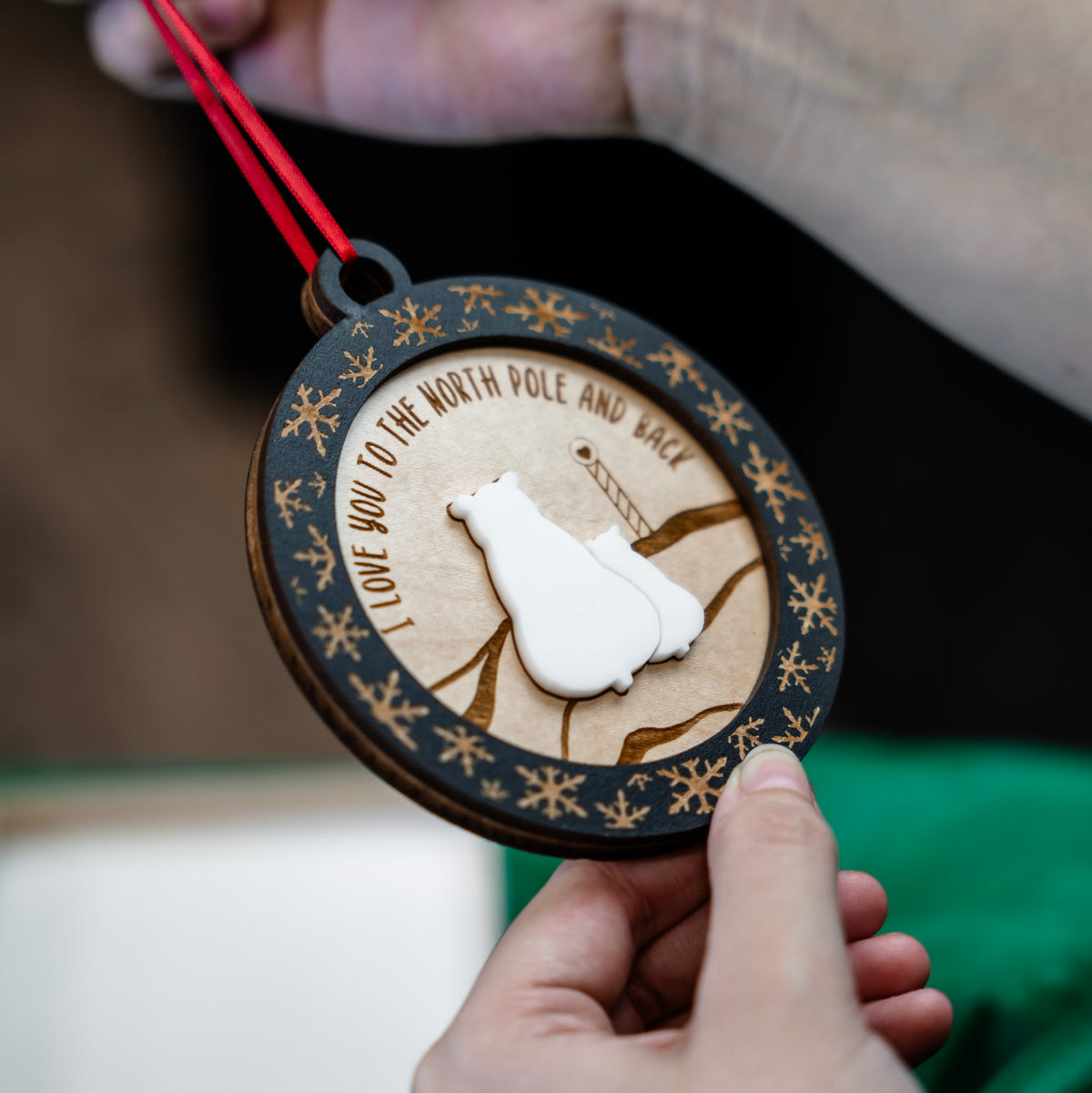A close-up of a round wooden ornament held by hands, featuring two polar bear silhouette in the center, laser-engraved snowflakes and text around the edge, and a red ribbon at the top.