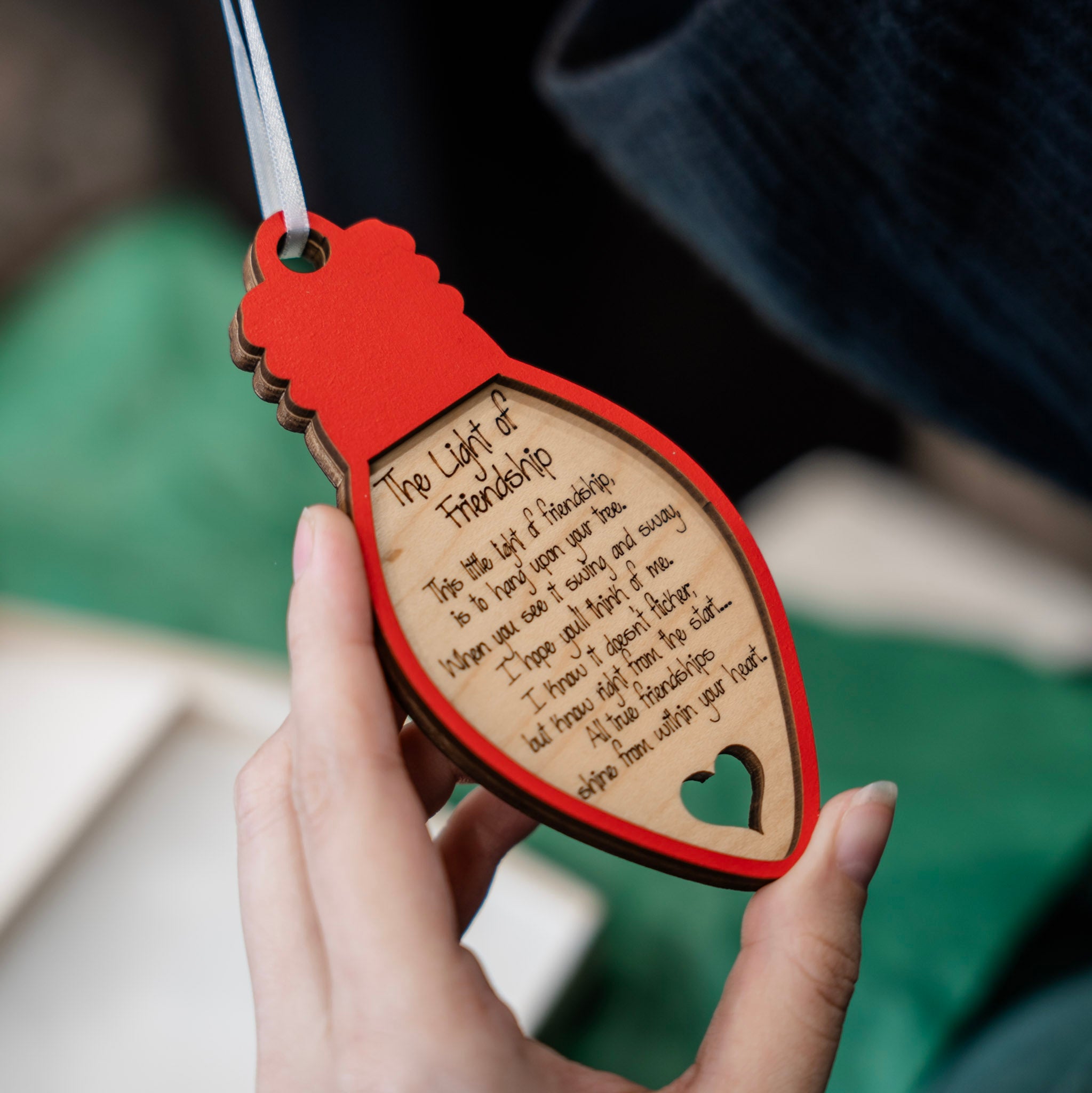 A hand holding a laser-engraved wooden lightbulb ornament with a red painted border and a white ribbon. The wooden lightbulb features a quote titled 'The Light of Friendship' and a small heart-shaped cutout.