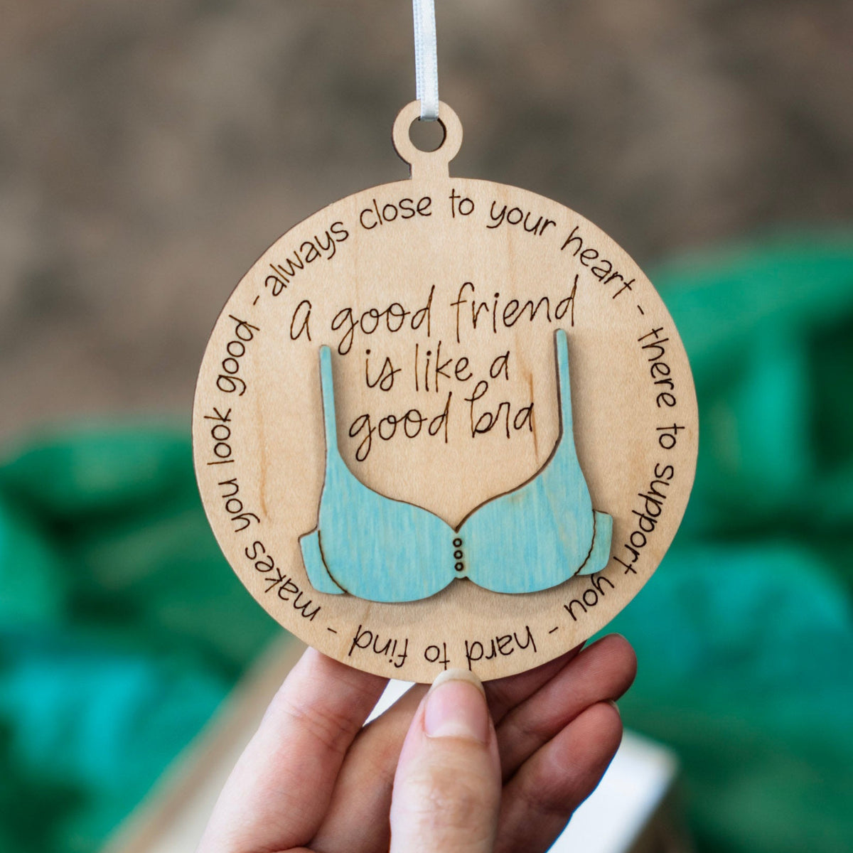 A hand holds a round wooden ornament engraved with a 3D bra and the quote 'a good friend is like a good bra' surrounded by supportive words. The background is softly blurred with green tissue paper and a neutral indoor setting.