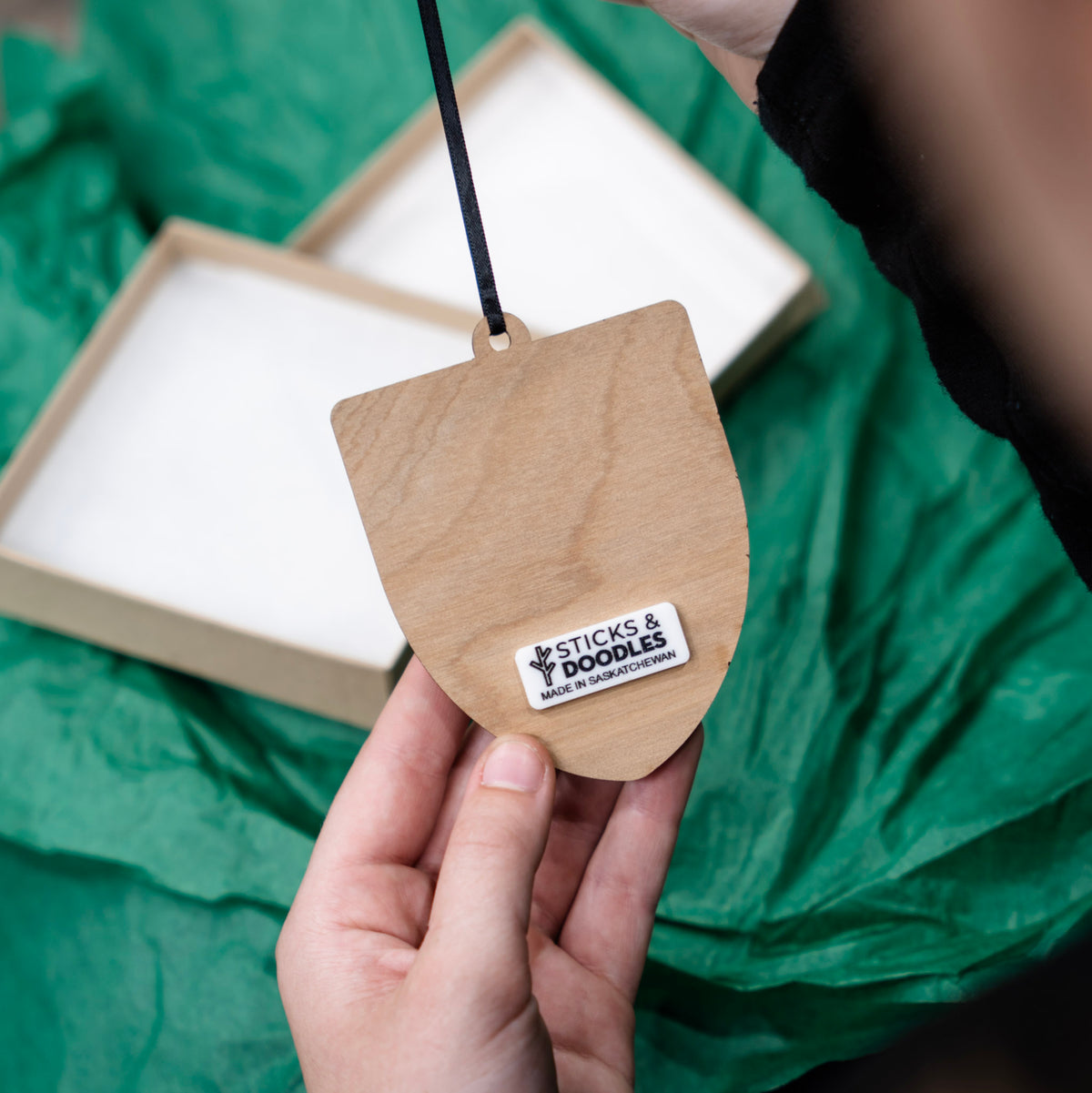 A hand holds a small natural wood shield-shaped University of Regina ornament with a black hanging string and a white 'STICKS & DOODLES' label. There is a box resting on top of the green tissue paper in the background.