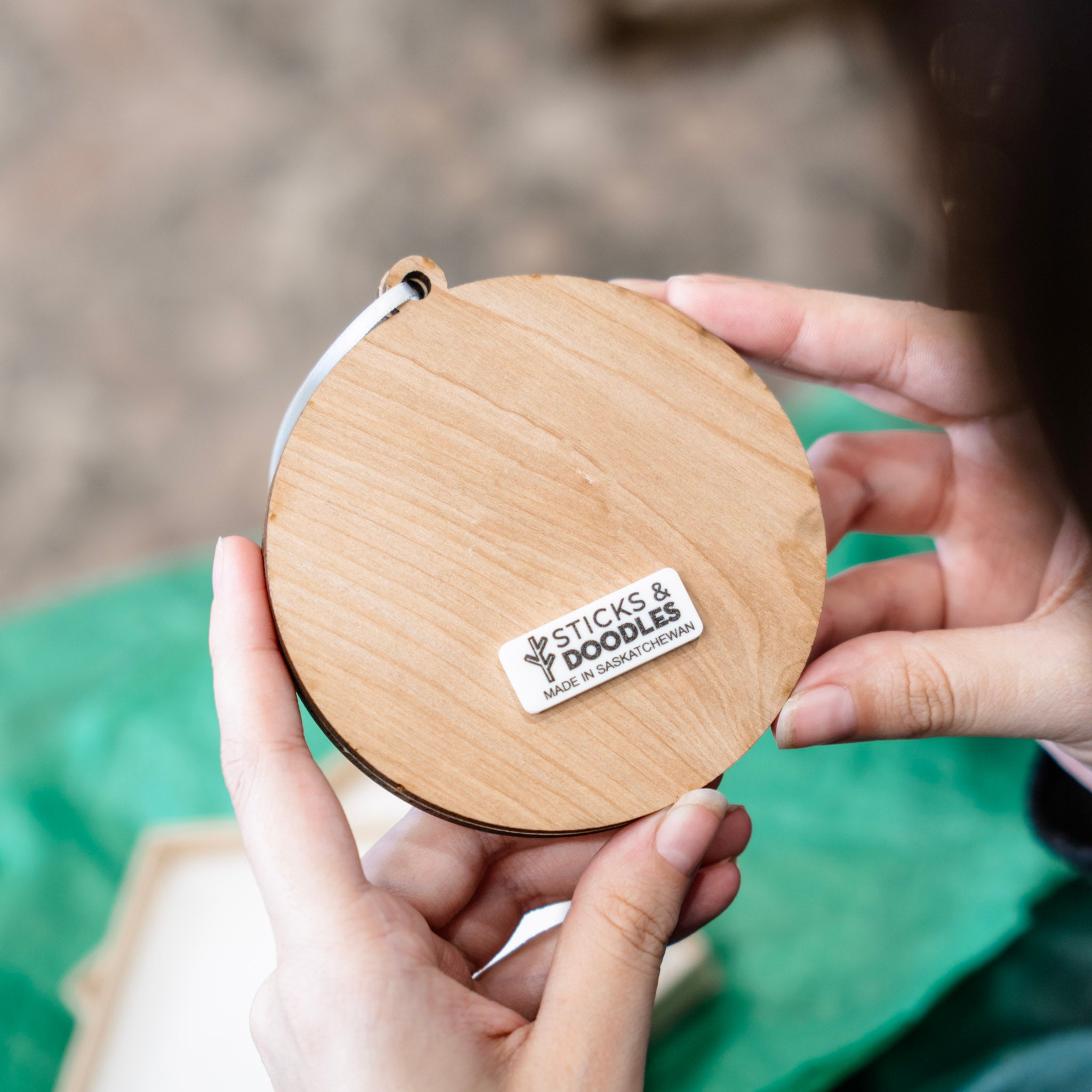 A person holding a round wooden ornament with a white tag that reads 'STICKS & DOODLES MADE IN SASKATCHEWAN'.