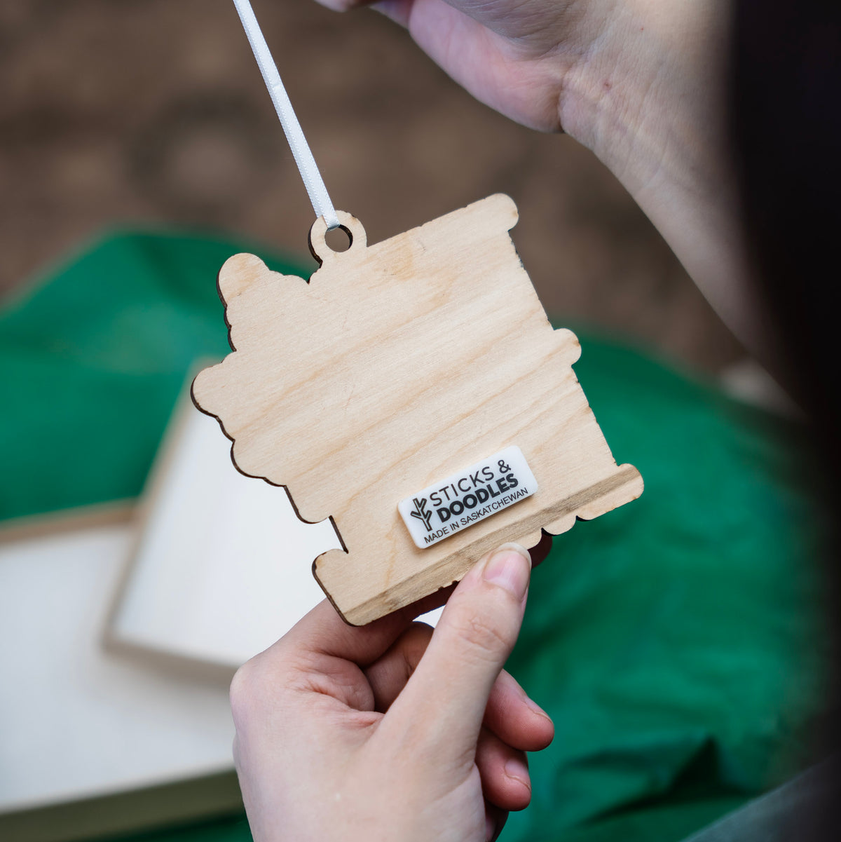 A close-up of a hand holding up the back of a wooden , with another hand holding the white ribbon at the top, set against a green fabric background.