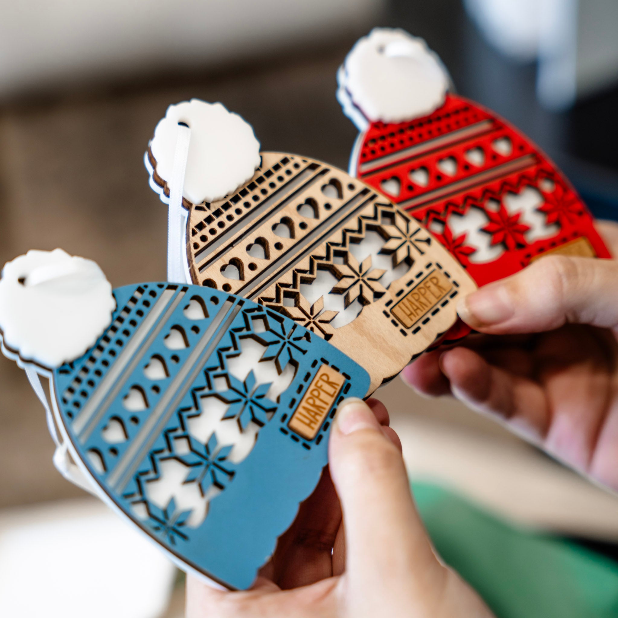 Three laser-cut wooden Christmas ornament shaped like knit winter Toques are held in two hands. The ornaments are in blue, natural wood, and red, each with a white faux-fur pom-pom and a small wooden tag that spells out 'Harper'.