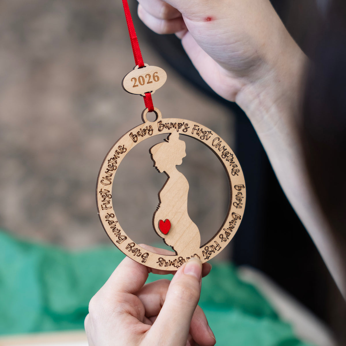 A close-up of a hand holding a personalized wooden baby bump ornament with a red ribbon. The ornament features a laser-cut silhouette of a pregnant belly with a small red heart, surrounded by engraved phrases, with a small wooden tag at the top engraved with '2026'.