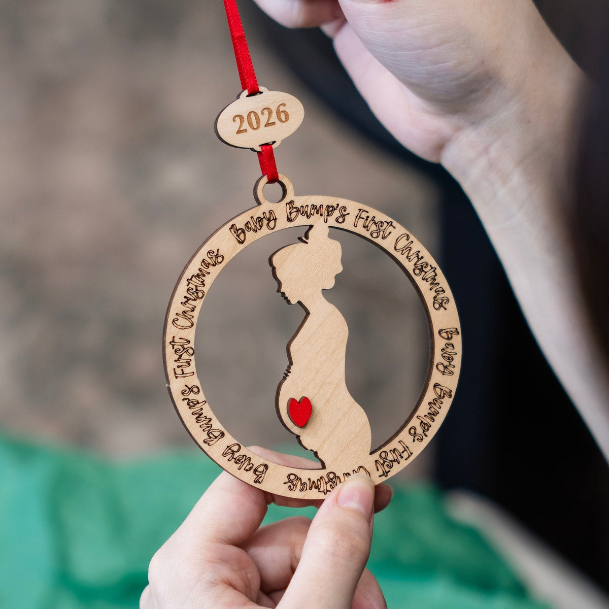 A close-up of a hand holding a personalized wooden baby bump ornament with a red ribbon. The ornament features a laser-cut silhouette of a pregnant belly with a small red heart, surrounded by engraved phrases, with a small wooden tag at the top engraved with '2026'.