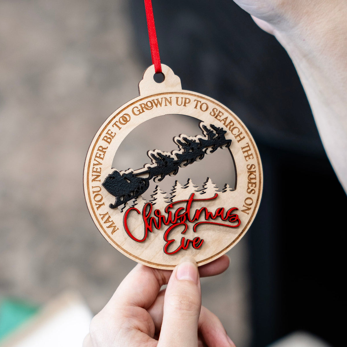 A personalized laser-engraved wooden Christmas Eve ornament held by hands, with a red ribbon, Santa's sleigh silhouette, pine trees, and the text 'May you never be too grown up to search the seas on Christmas Eve'.