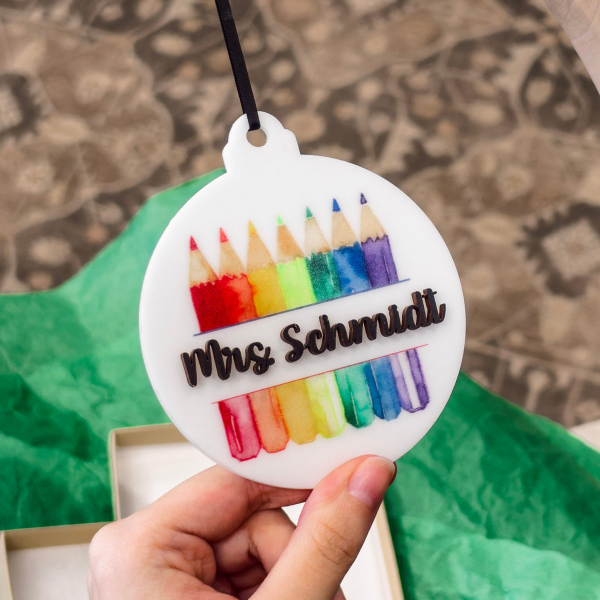 A hand holds a round white ornament featuring colorful pencil illustrations and the name 'Mrs. Schmidt' in black script, with a black hanging ribbon above it.