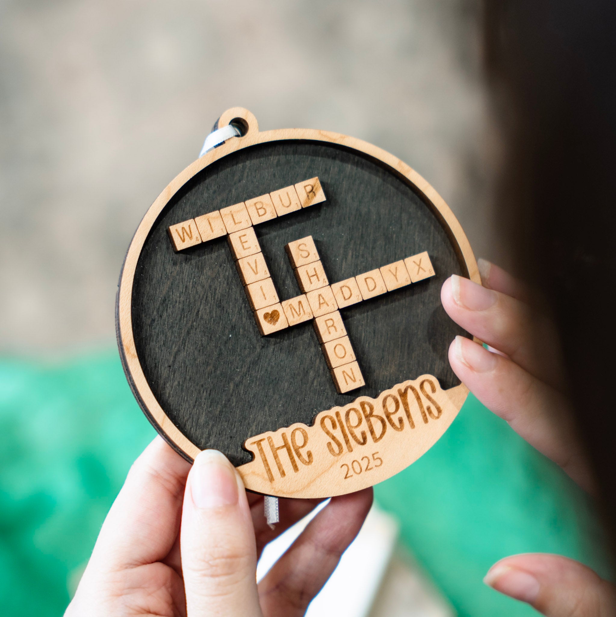 A personalized round wooden ornament held by two hands, featuring a dark wood center with a crossword-style arrangement of Scrabble tiles with four different names, plus the engraved text 'The Siebens' and '2025' on a light wood border.