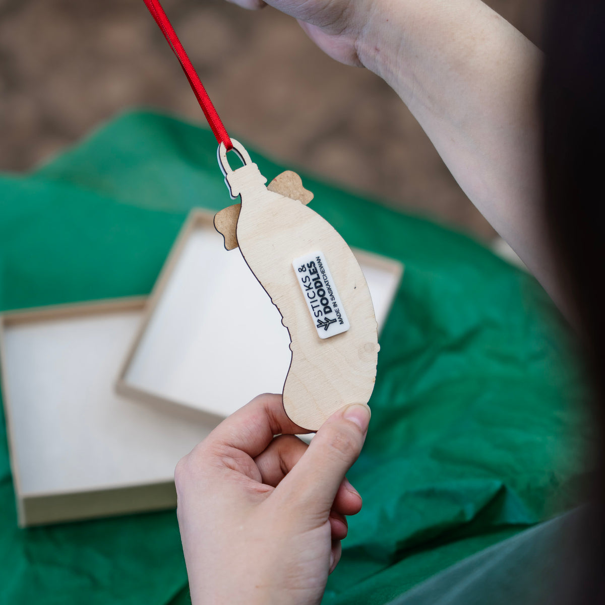 A hand holds the back of a pickle ornament to show the white tag that reads Sticks & Doodles Made in Saskatchewan. Hanging from a red ribbon.