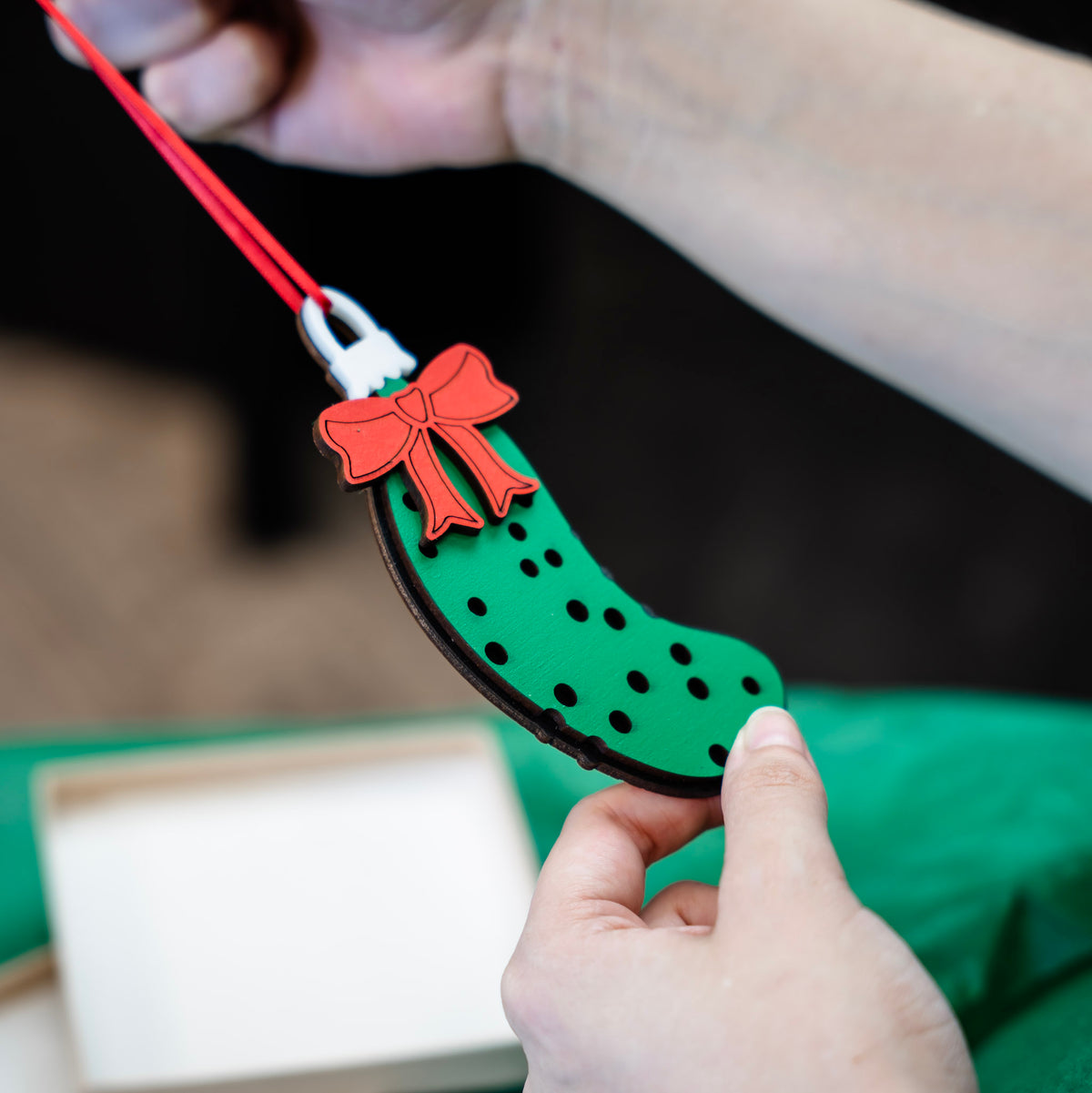 A close-up of a person holding a green Christmas pickle ornament with a red bow and red hanging string against a dark, blurred background.