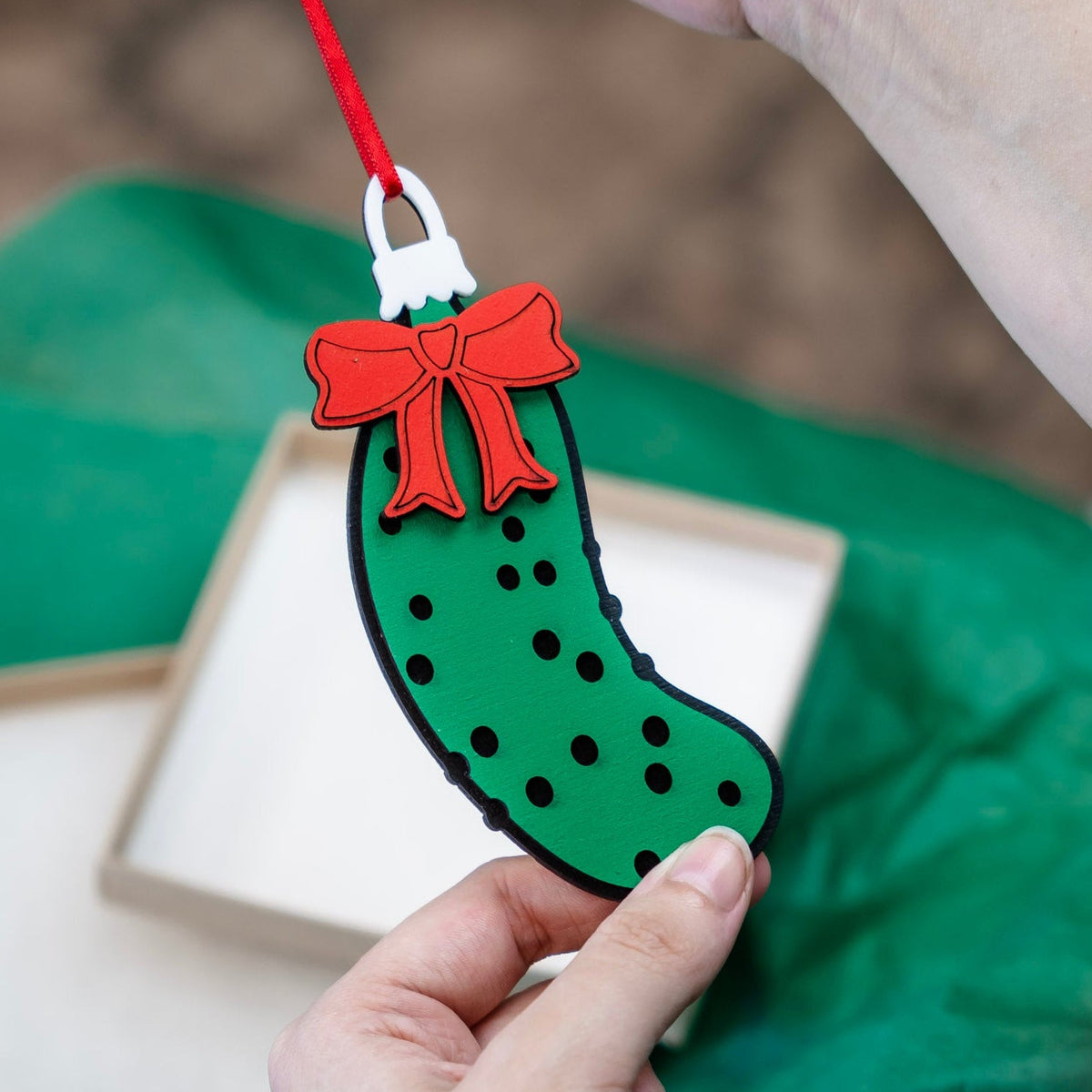 A person holds a Pickle Christmas ornament with a red bow, hanging a red string above a gift box on green tissue paper.