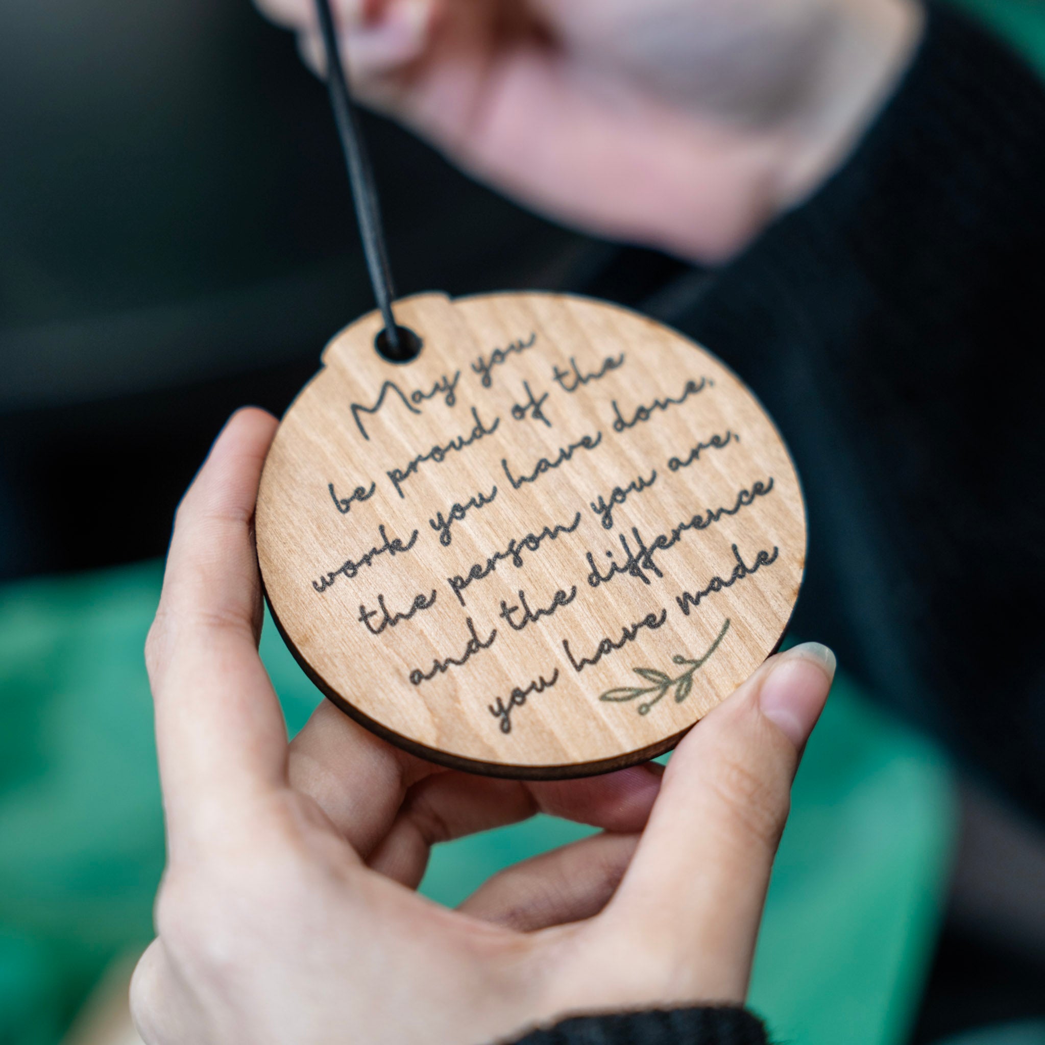 A person holding a round wooden engraved plaque with a black hanging cord. The plaque features a quote in cursive script and is shown close-up against a blurred background.