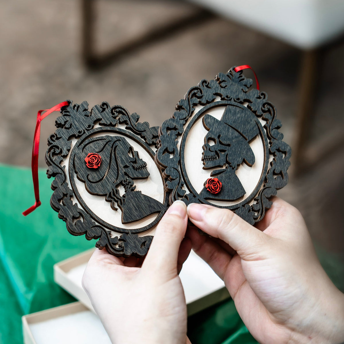 A person holds two ornate black wooden Christmas ornaments with red ribbons and small red rose details, showing skull and profile cutout designs. A gift box and green wrapping paper are visible in the background.