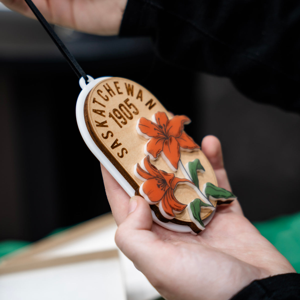 A close up of a hand holding a three layer wooden and acrylic ornament engraved with 'SASKATCHEWAN 1905' and two orange lilies on top . An open gift box and green wrapping paper are visible in the background.