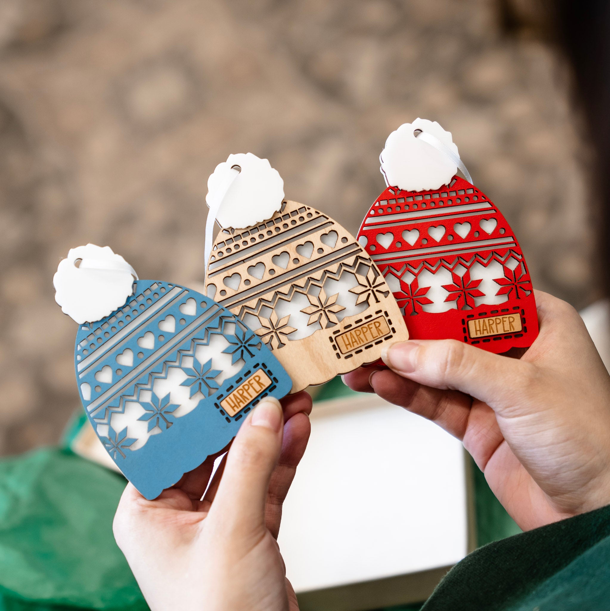 Three laser-cut wooden Christmas ornament shaped like knit winter Toques are held in two hands. The ornaments are in blue, natural wood, and red, each with a white faux-fur pom-pom and a small wooden tag that spells out 'Harper'.