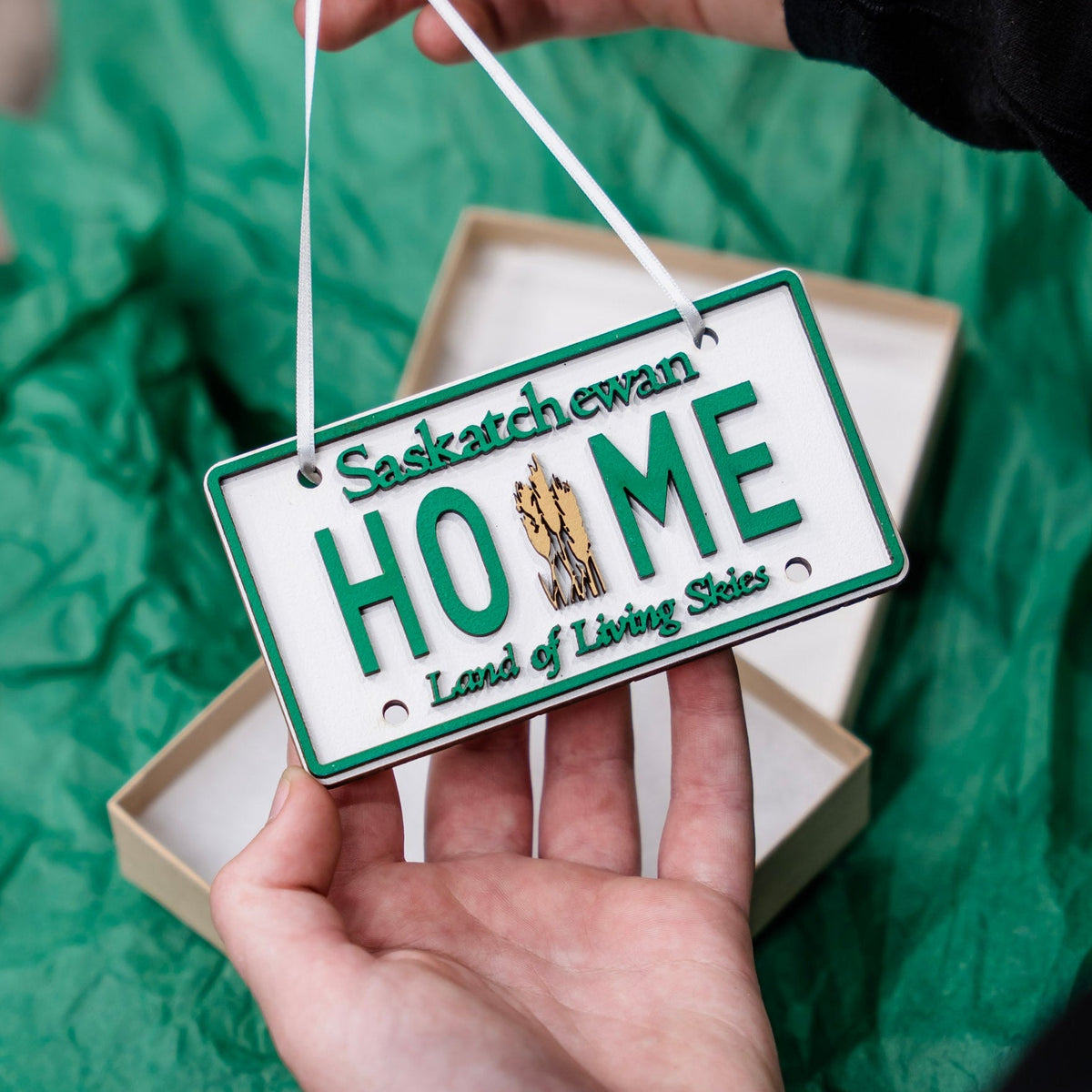 A hand holds a white and green license plate ornament shaped like a Saskatchewan license plate, with the word 'HOME' in the center and a small tree graphic. The ornament hangs from a white ribbon, with a beige gift box and crumpled green paper in the background.