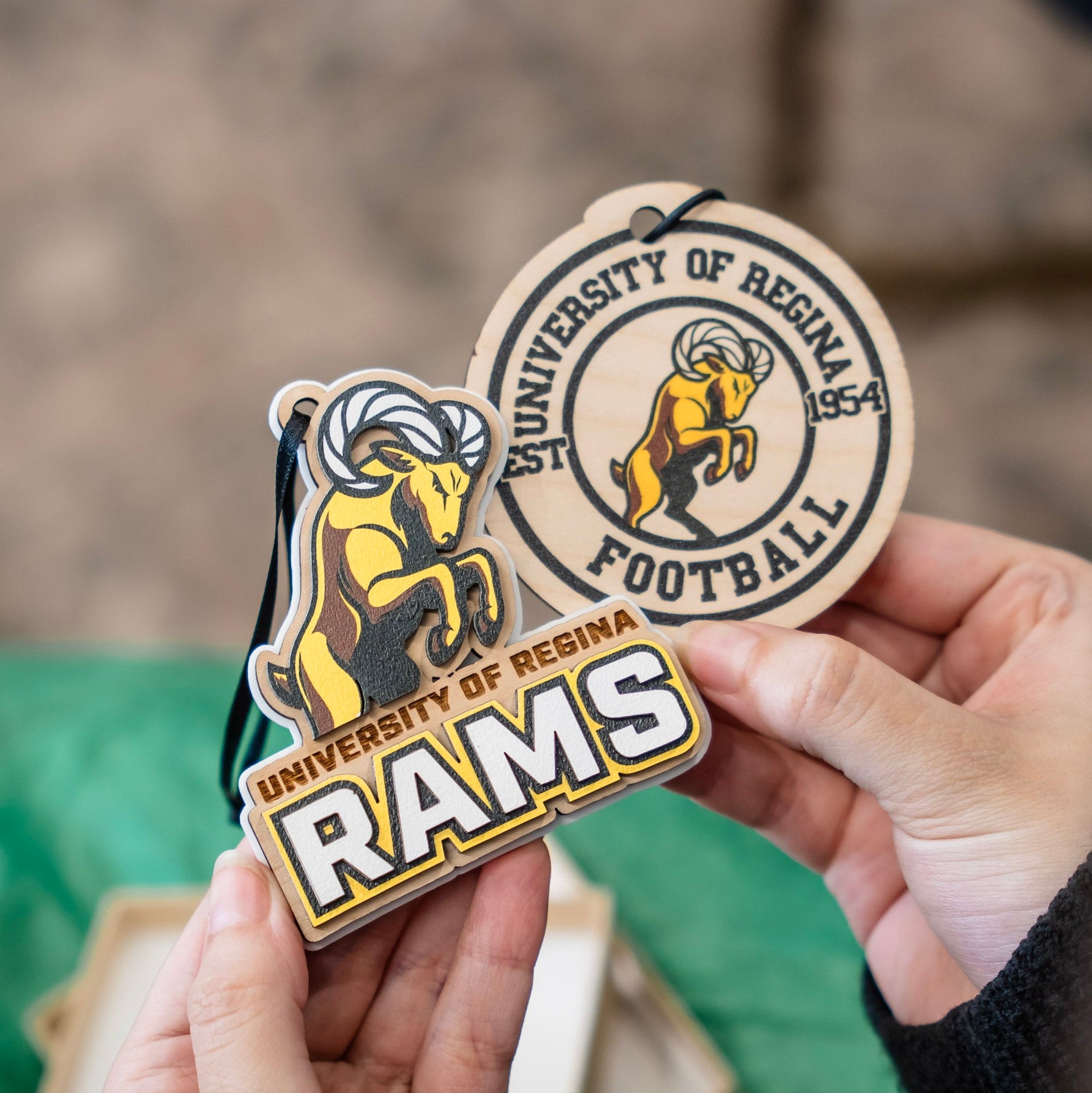 Two University of Regina Rams ornament and car charm held in hands: a round wooden football tag and a matching wooden ram-shaped ornament, both featuring the yellow ram logo and school name.