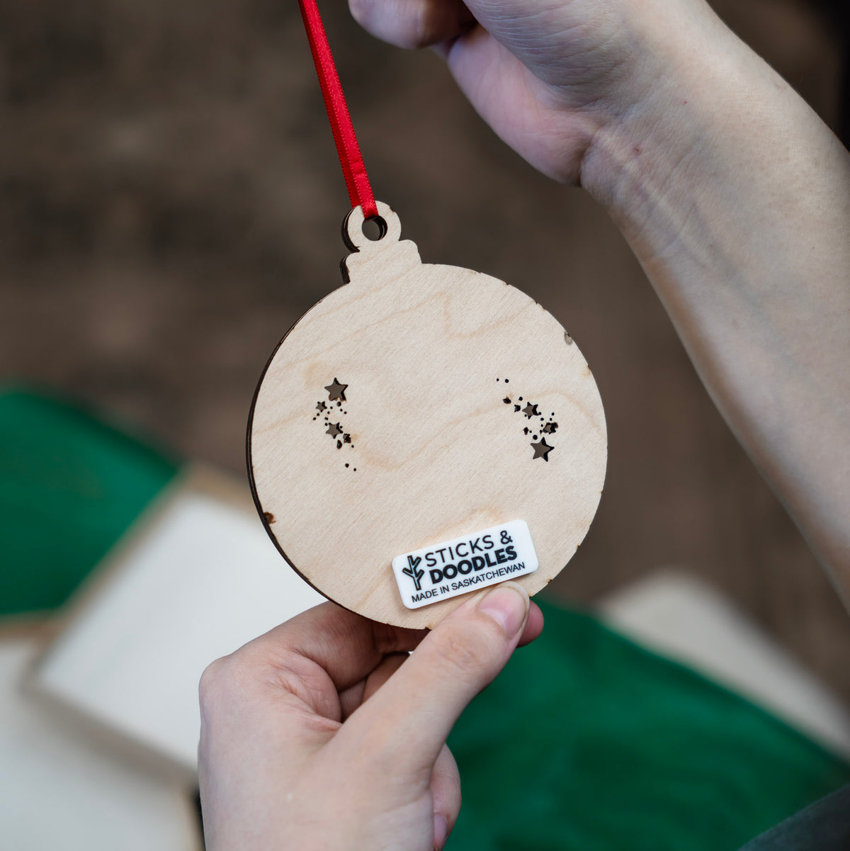 A person holds the back of the Santa in a Chimney ornament, showing the star cutout and red ribbon suspending the wood ornament. There is a acrylic tag on the back that reads STICKS & DOODLES MADE IN SASKATCHEWAN.