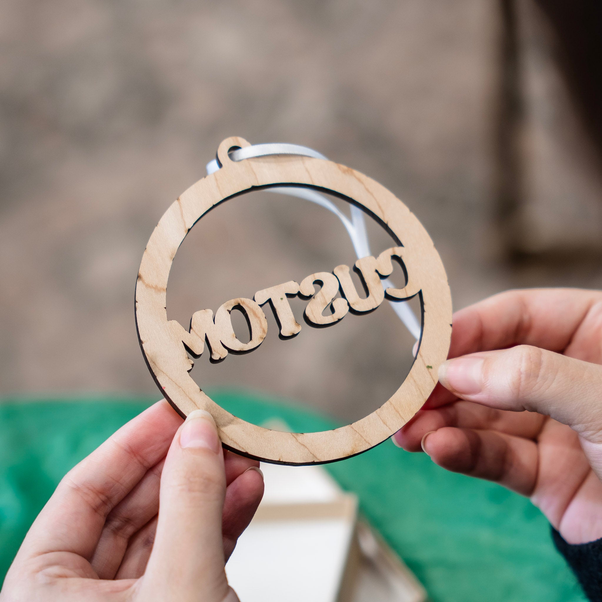 A person holds the back of the circular wooden ornament with the word 'CUSTOM' cut out in the center, showing a white hanging ribbon. The background is softly blurred with a green surface below.