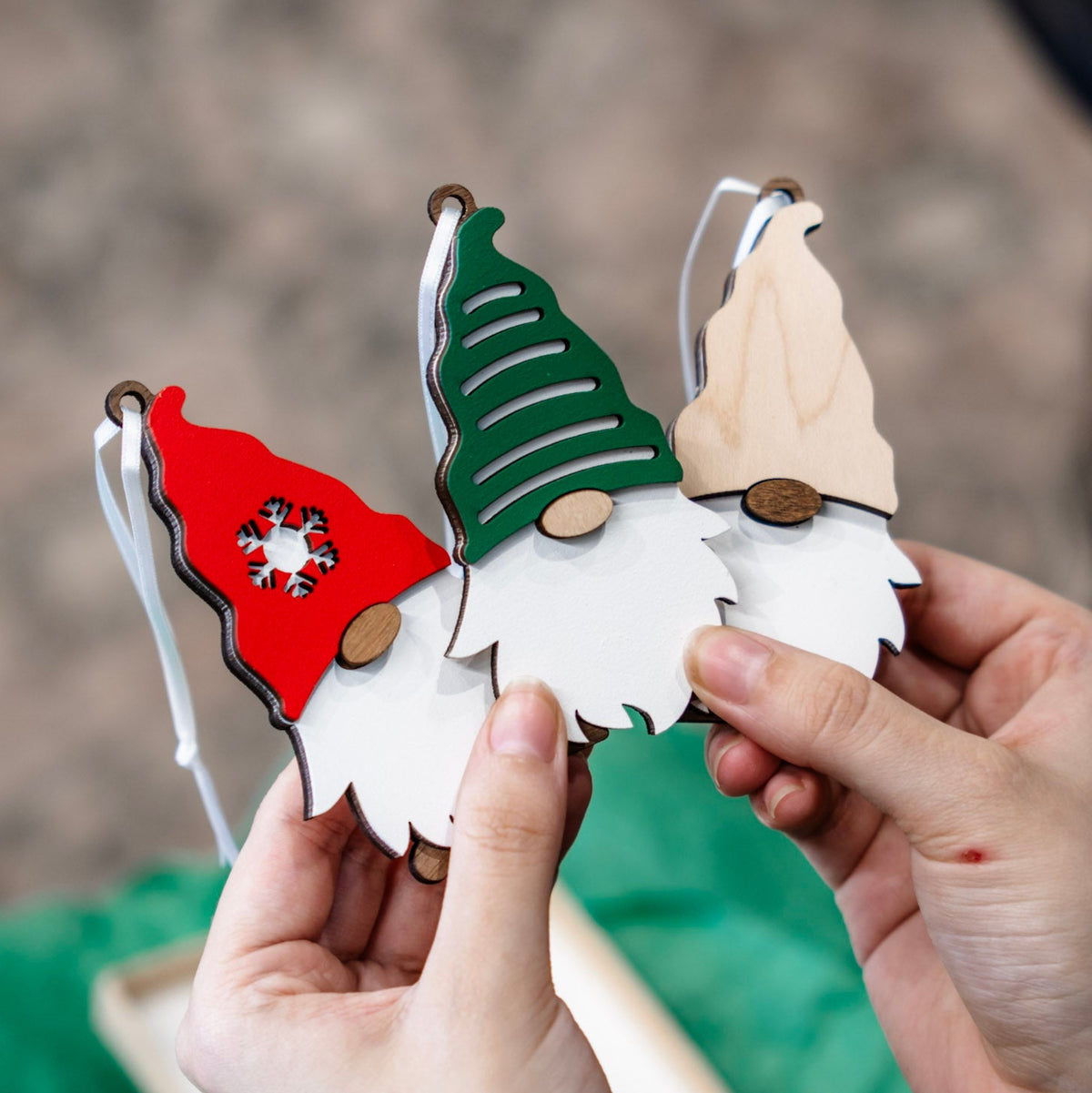 Three wooden gnome ornaments with white beards are held in two hands against a blurred outdoor background. One ornament has a red hat with a white snowflake, one has a green hat, and one has a natural wood hat.