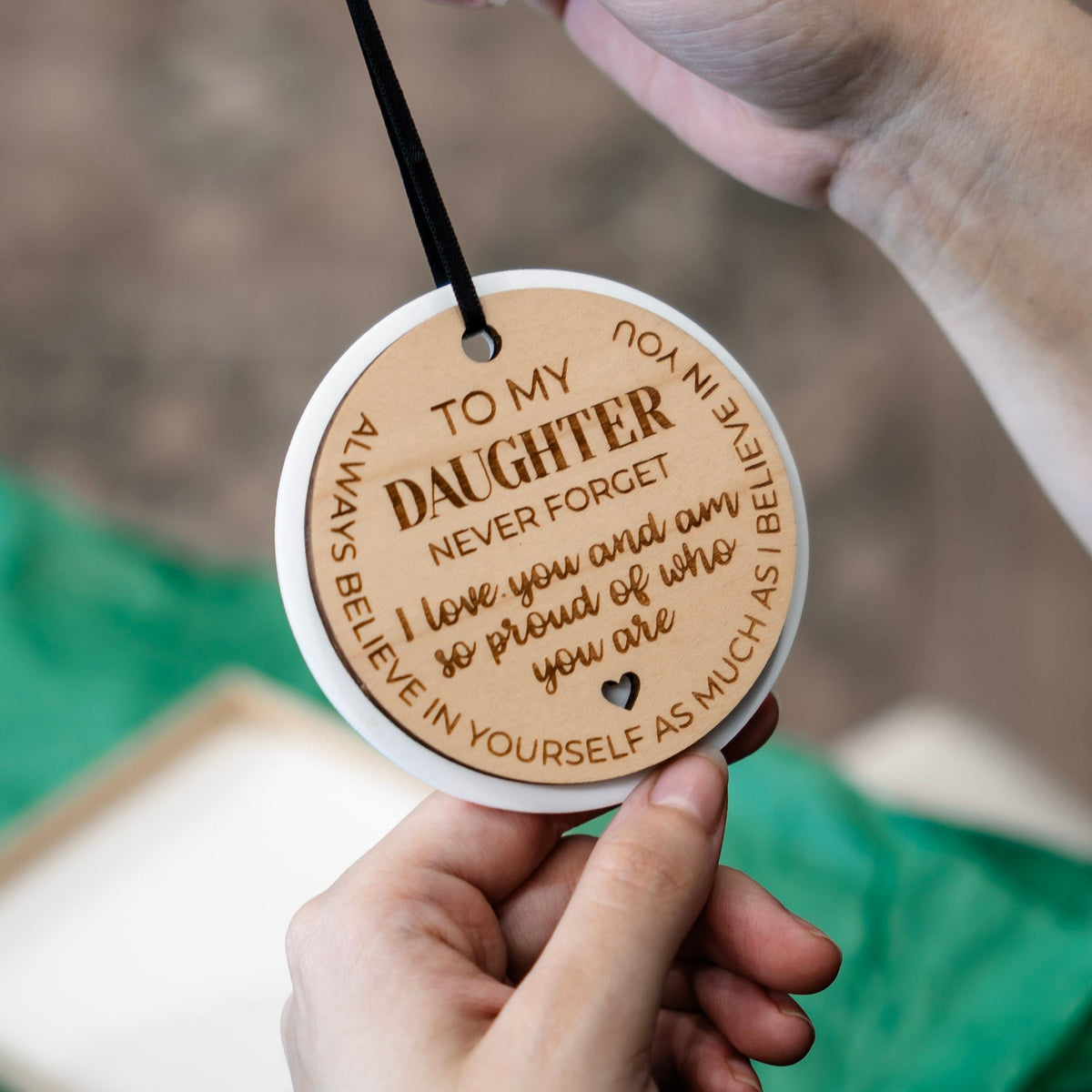 A close-up of a person holding a round wooden ornament with engraved text for a daughter. The ornament is light brown wood with engraved dark brown lettering and a black ribbon, shown against a blurred background with green tissue paper.