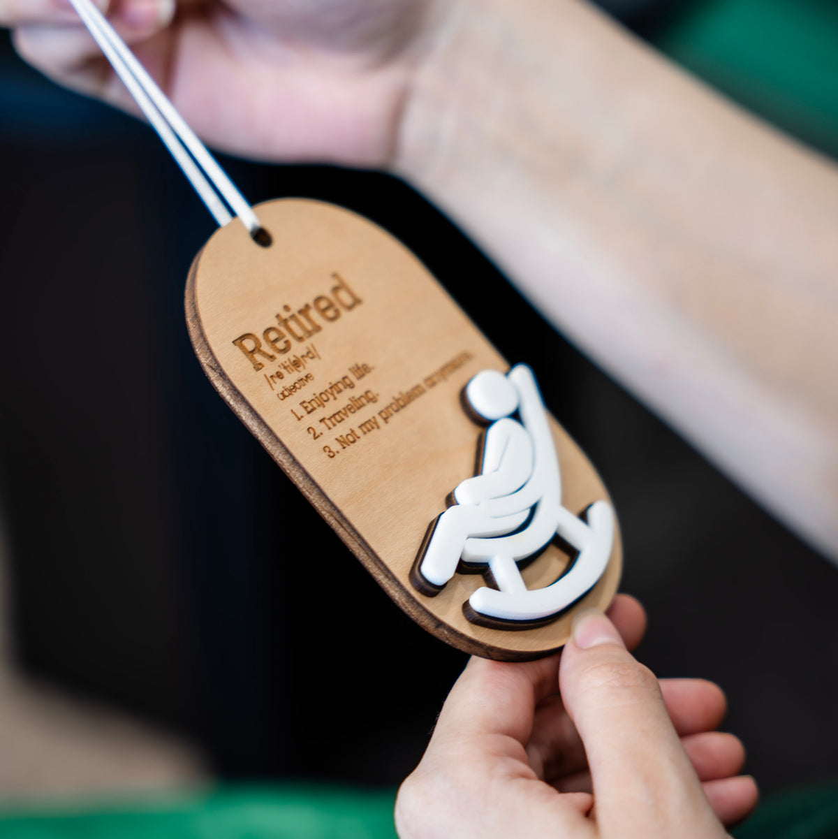 A person holds a wooden ornament with a white ribbon. The tag is engraved with the word 'Retired' and a definition, with a white silhouette of a person sitting in a rocking chair.