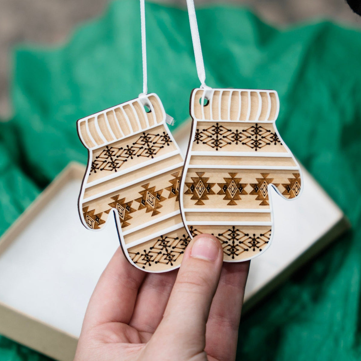 A hand holds two wooden Christmas ornament shaped like mittens with a laser-engraved and scored geometric pattern. The ornaments are suspended by white ribbon, with a gift box and bright green tissue paper in the background.