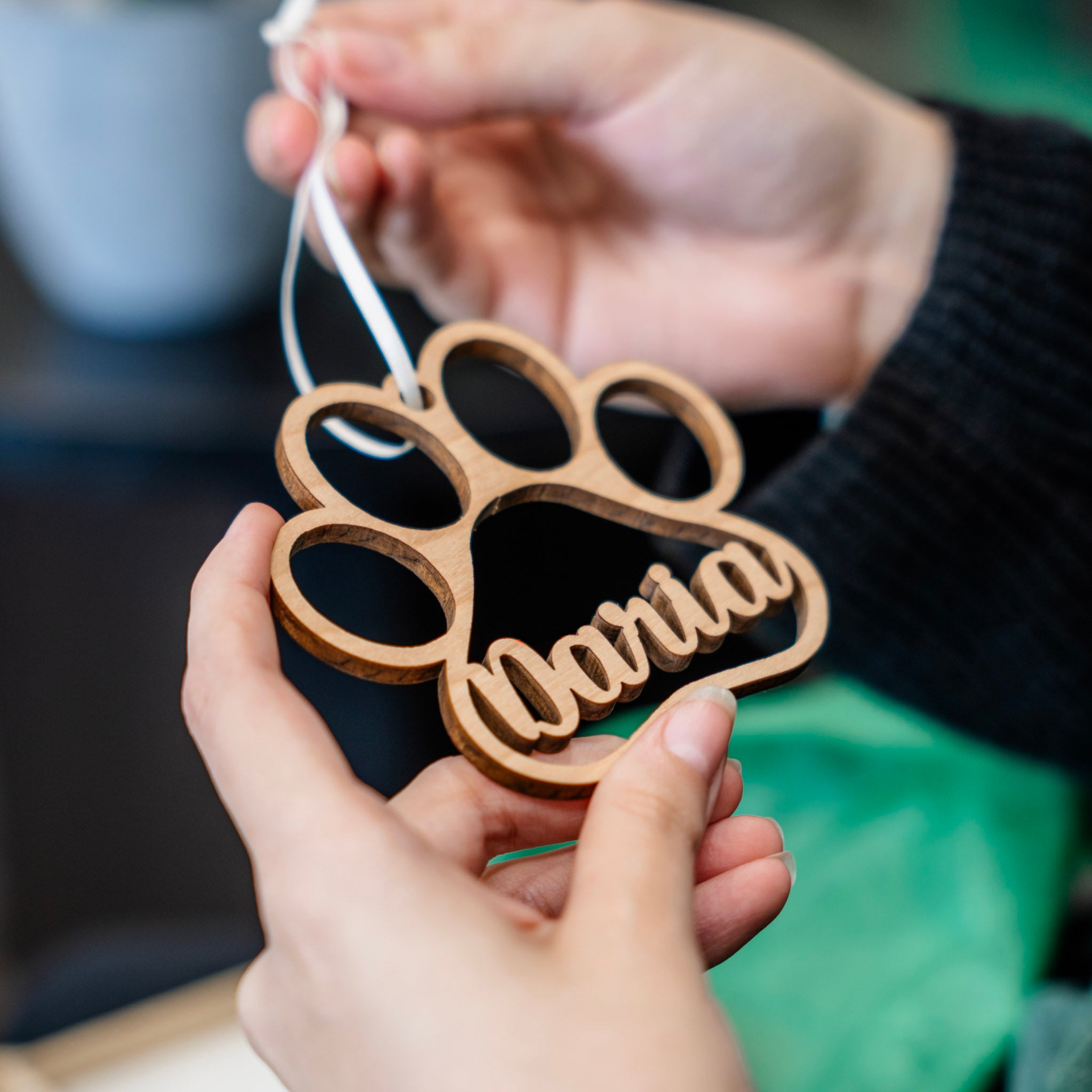 A wooden paw-shaped ornament with the name 'Daria' cut into the wood, held in two hands. The ornament is made of light natural wood and hangs from a white ribbon.