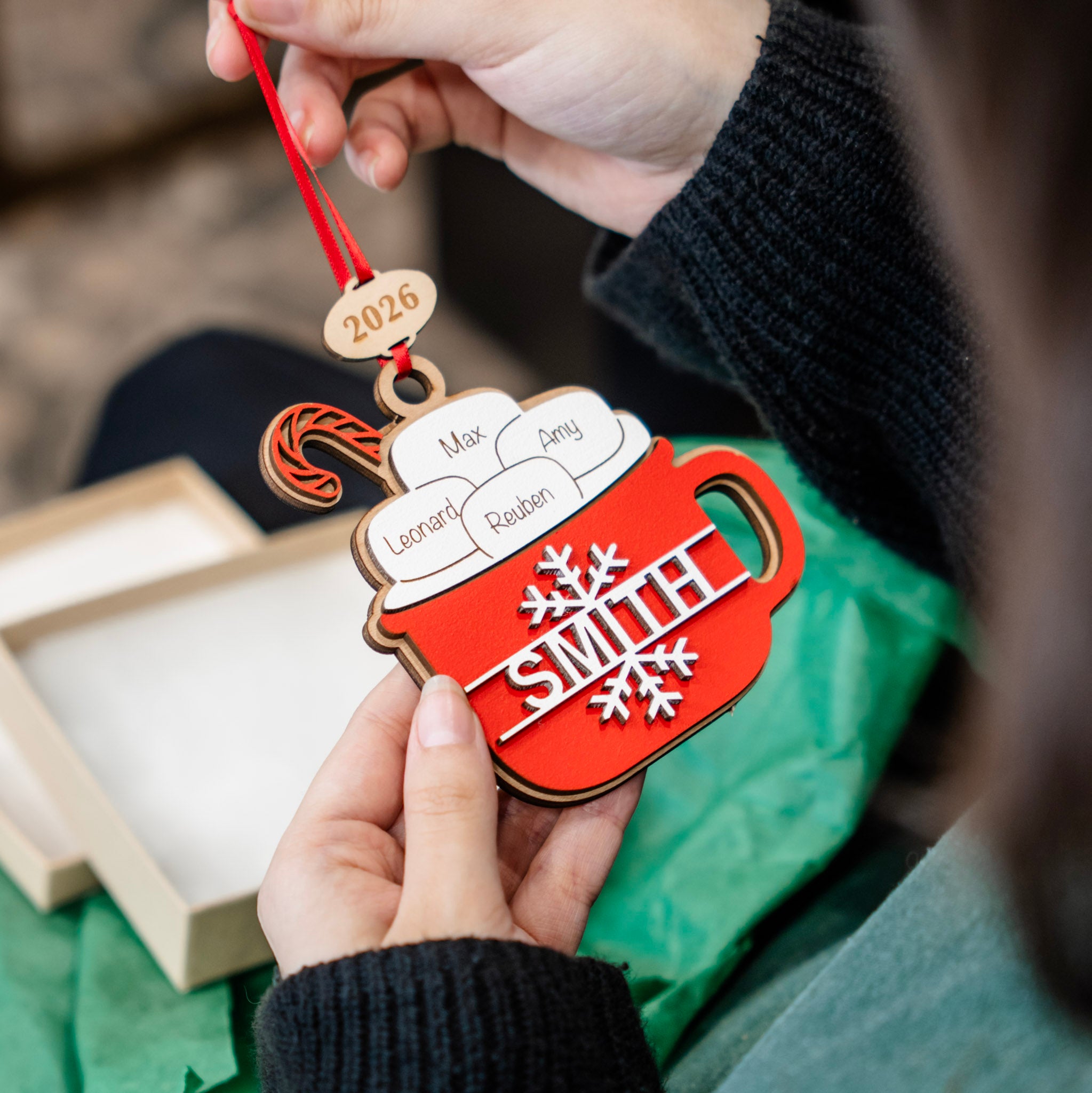 A person holds a personalized wooden mug Christmas ornament shaped like a red coffee mug with white steam, custom names on the steam, a snowflake and the name 'SMITH' on the mug, and a 2026 wooden tag hanging from a red string.